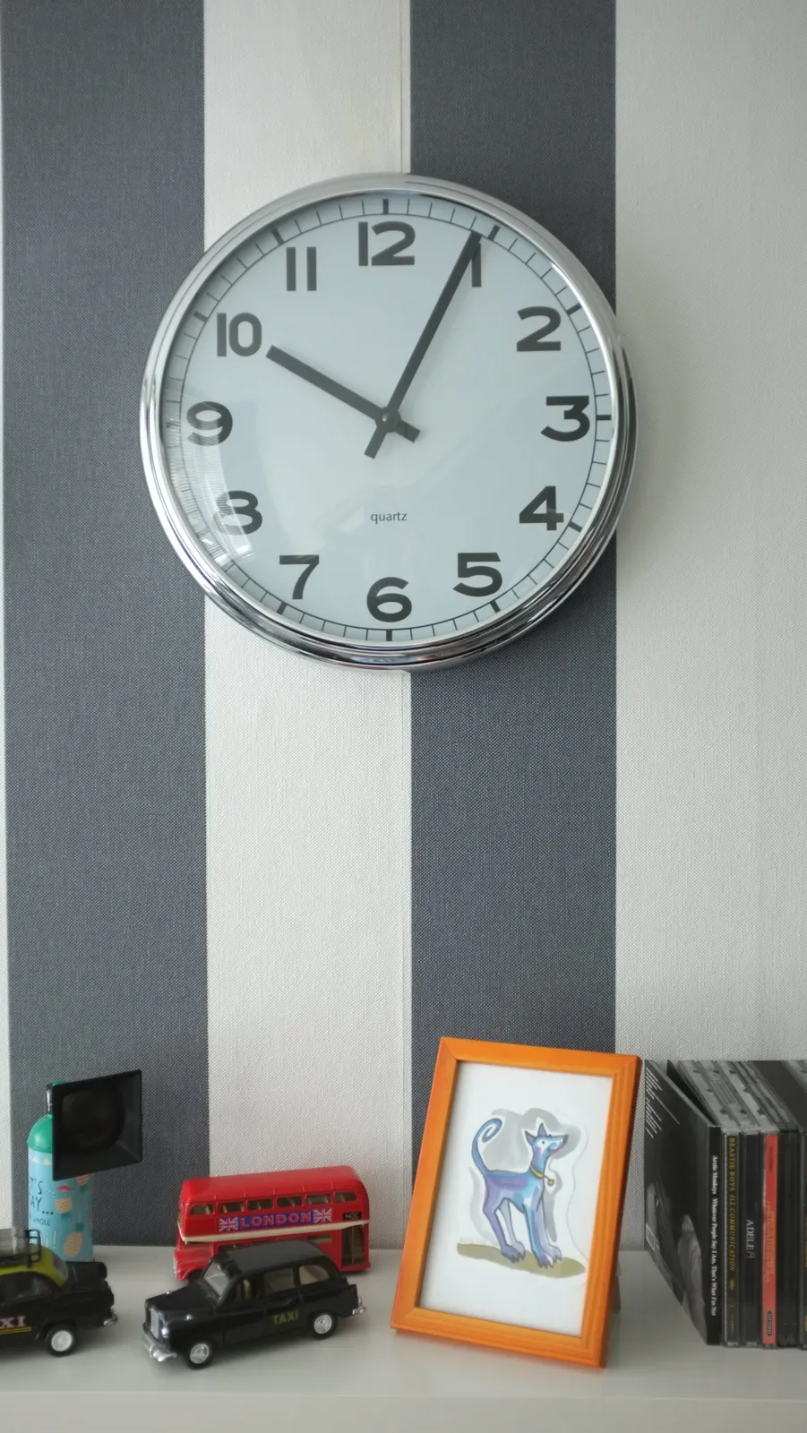 A clock on a striped wall. It points at 4 minutes past 10. Below the clock there are some CDs and some other stuff.