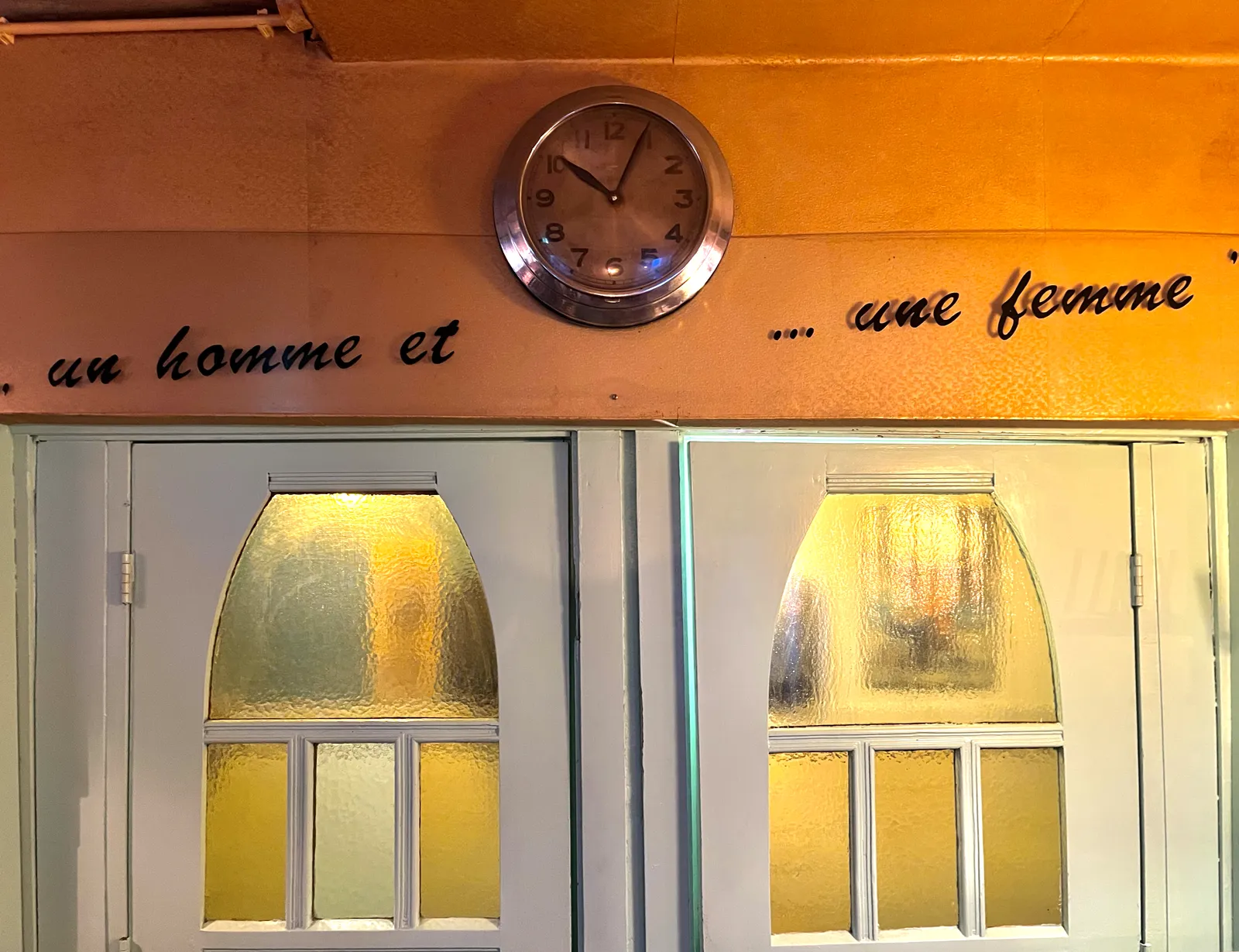 Above two doors it says, in French, un homme et … une femme. Above this sentence there’s a wall clock that points at 4 minutes past 10.