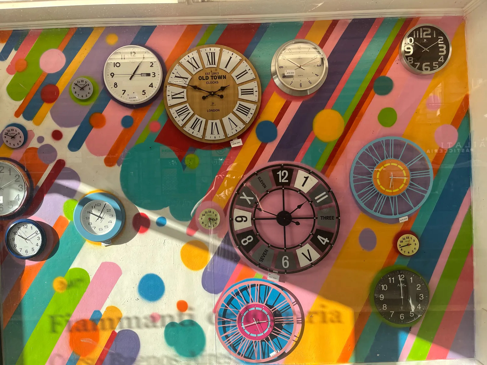 A very, very colourful shopping window on a sunny day, with lots of clocks in it. There’s a clock that points at 5 past one. There are a few clocks that point somewhere between 12 minutes to 2 and 10 minutes to 2. There’s a clock that points at 42 minutes past 2, and one that points at 12 minutes to 3. There’s also a clock that points at 28 minutes past 3. And there’s one that points at 16 minutes past 6. Then there’s one that points at 17 minutes to 9, there’s one that points at 5 past 10, there are a few clocks that point somewhere between 9 minutes past and 11 minutes past 10, and finally there’s this one clock that points at one minute to 12.