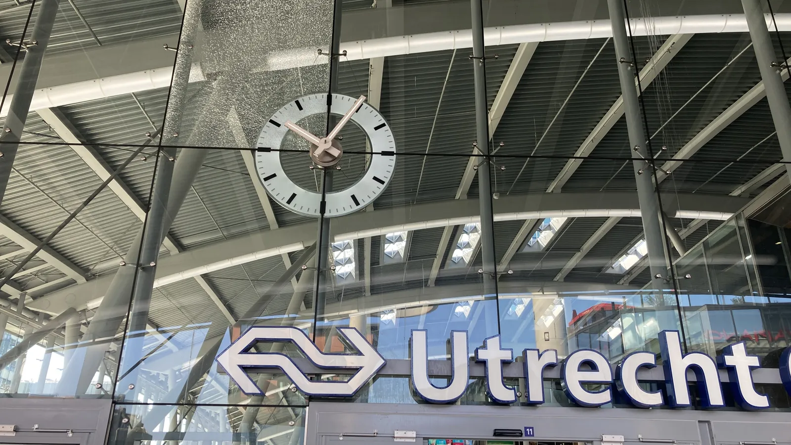 The large clock in the glass facade of Utrecht central station. One of the glass panes is shattered. The clock points at 5 past 10.