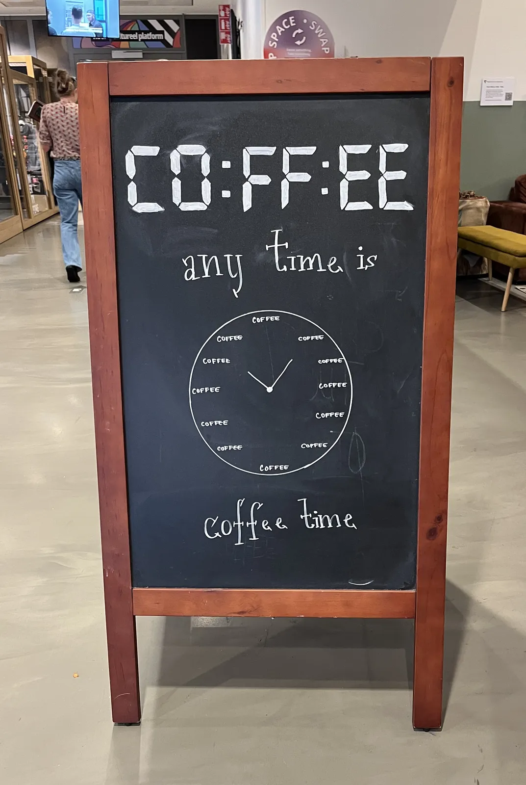 On a blackboard sign an analogue clock is drawn. Above this clock, in segmented digital letters, those use by alarm clocks, it says Coffee. Around the analogue clock it says: Any time is coffee time. All the hour indicators on the clock are made of the word Coffee. This coffee clock points at five past ten.