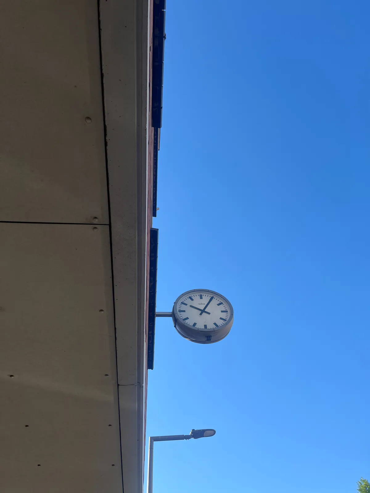 Looking up, from below a balcony, at a clock that’s attached to the building. The sky is bright blue. The clock points at 5 past ten
