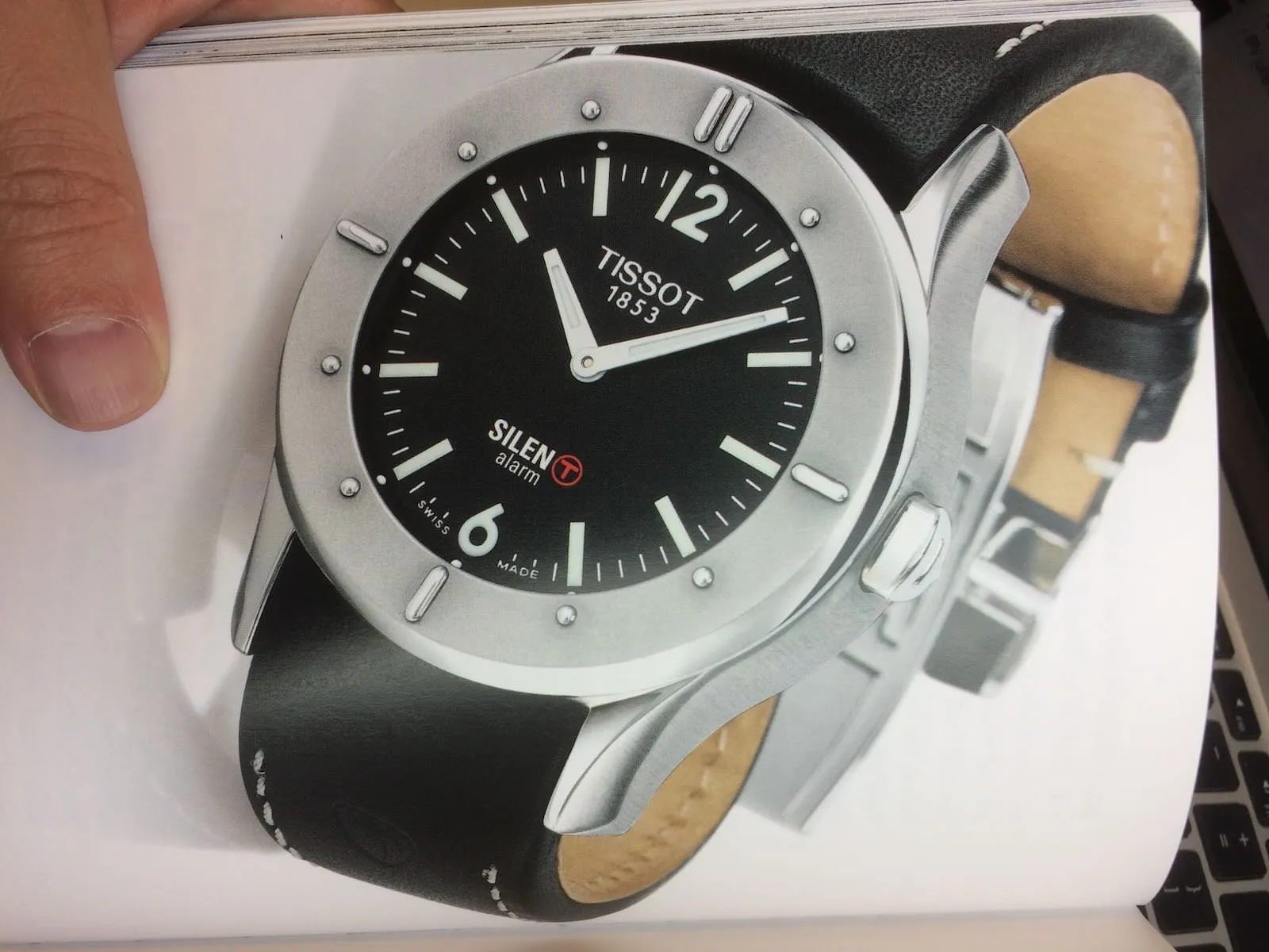A hand that holds a magazine with a photo of a watch on it. On this watch it is seven past ten.