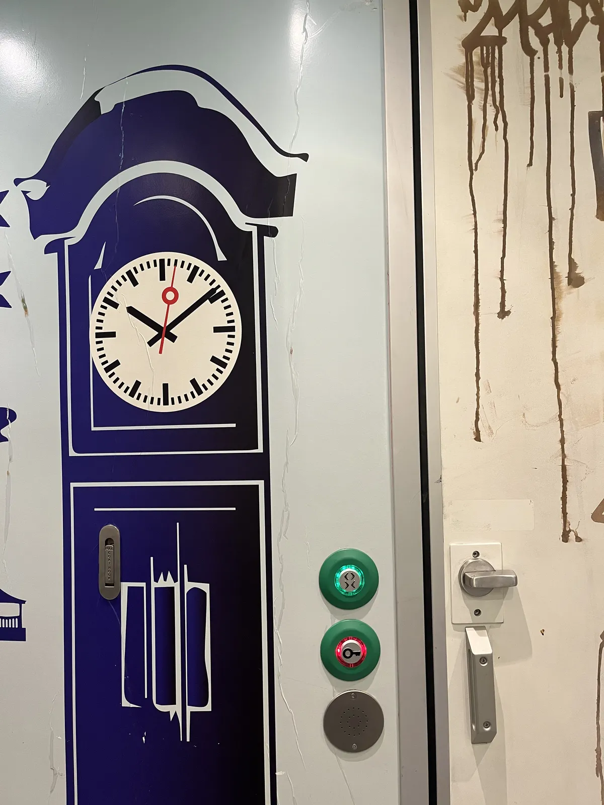 A large grandfather’s clock is painted on a wall. The face of the clock resembles the classical, modernist Dutch station clocks. Next to this clock are two buttons, one is lit green, the other red. The red one has a symbol of a key on it. Next to these buttons there’s a door handle and a knob to lock the door. There’s some dripping graffiti painted on this door. The clock points at 9 minutes past 10.