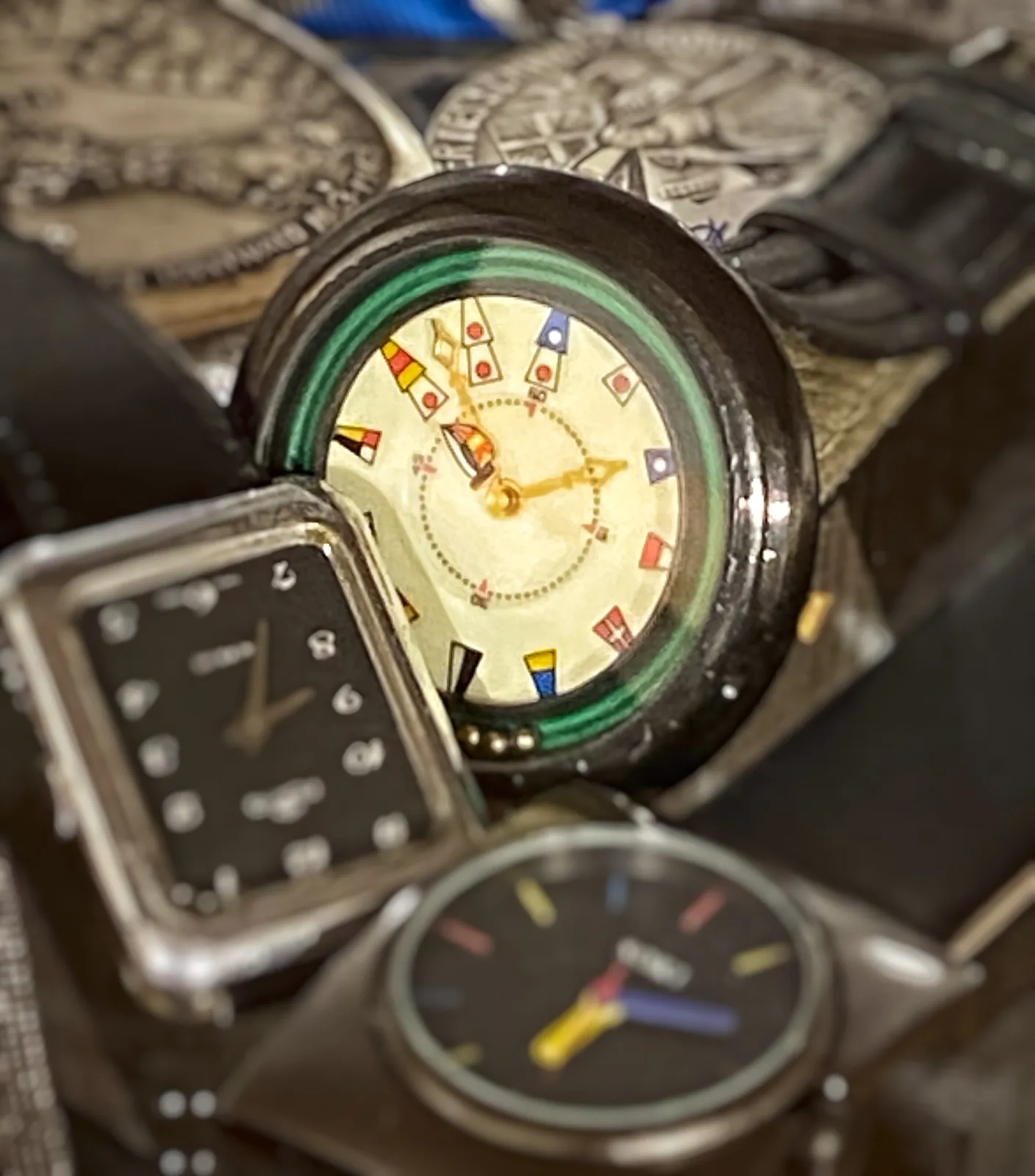A pile of watches. The one that’s in focus is hard to read. Instead of numbers for each hour it shows flags. Luckily there are a few numbers for printed for the minutes. Thanks to those we can now understand that it points at 9 minutes past 10.