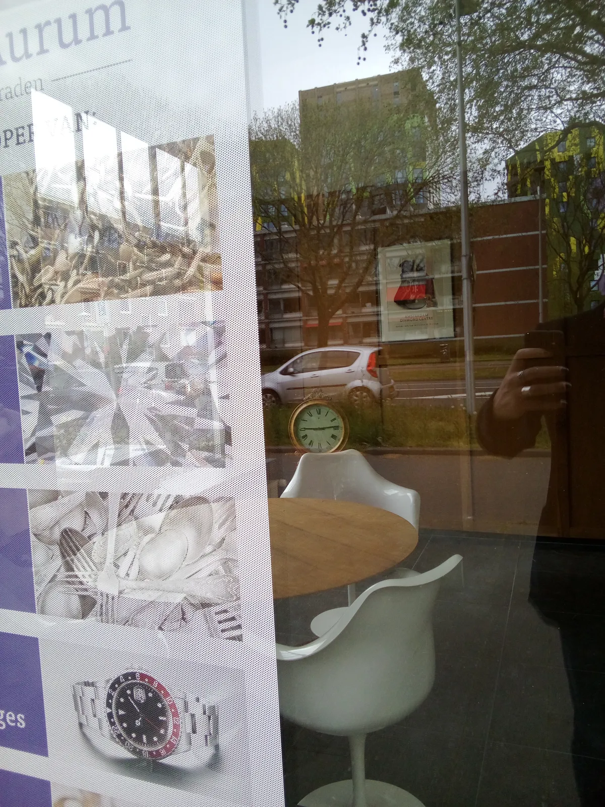 Peeking through a window. In the reflection the photographer can be seen, holding a phone, with a suburban city scape in the background. Right behind the window there is a big poster with some images of luxurious things on it, among others a wrist watch that points at 9 minutes past ten. In the background, inside the building, there’s a large clock that points at 14 minutes past nine. 