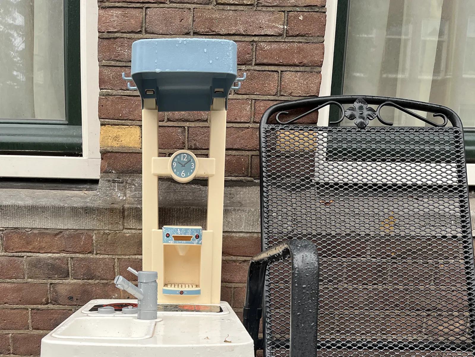 Outside, next to a black metal chair, there’s this toy kitchen with a sticker of a clock on it that points at 9 minutes past ten.
