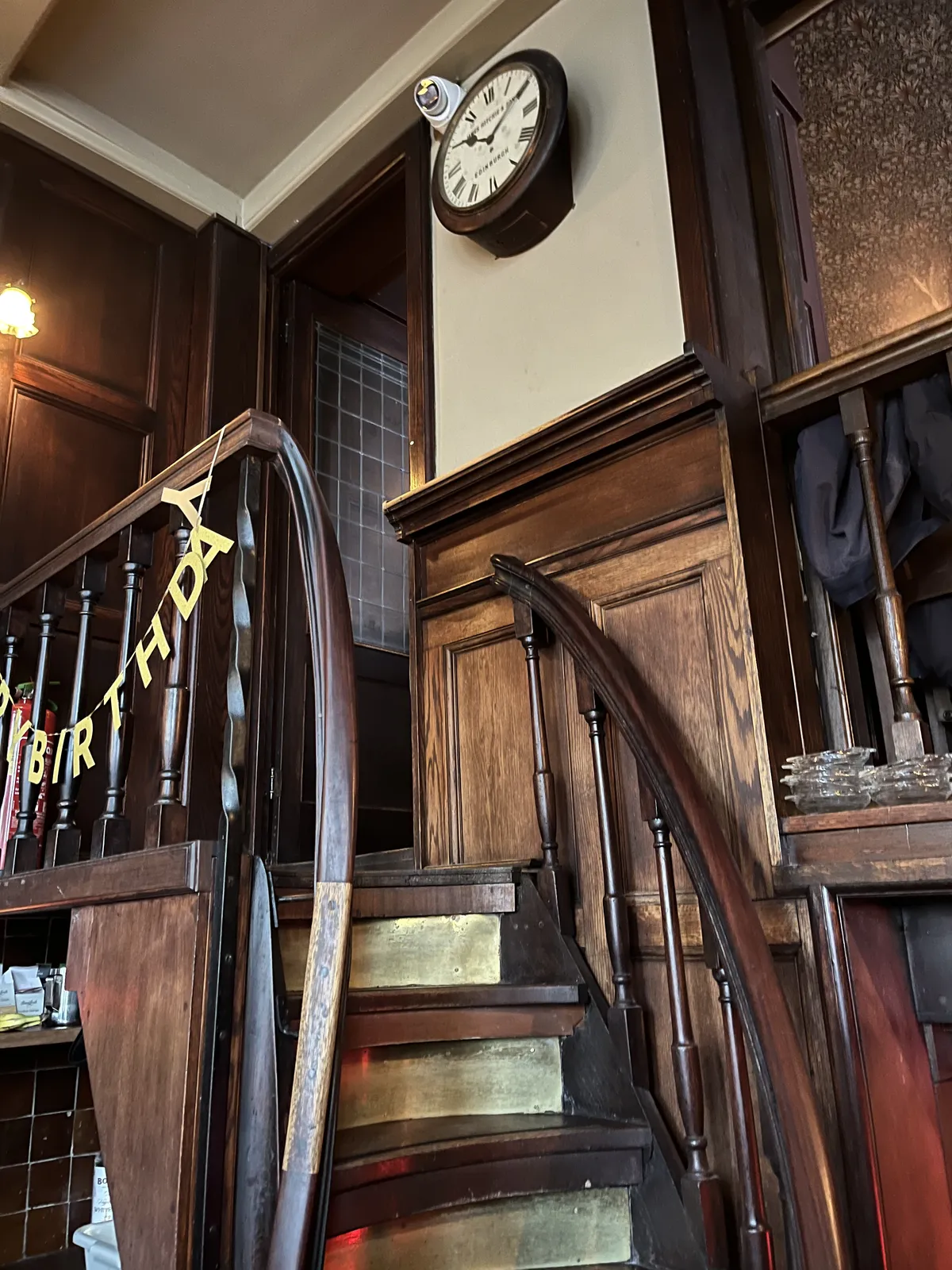 Looking up at some steep wooden stairs. On the wall next to the stairs there’s a clock that points at 10 past 10.
