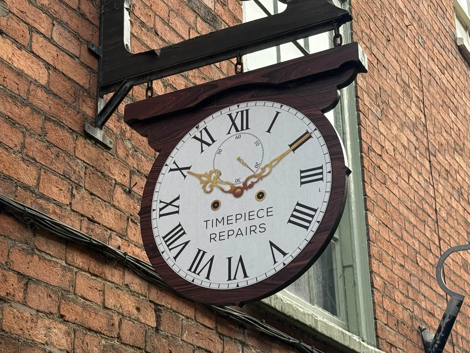 A fake clock, attached to a brick building. It says Timepiece repair on the face of this fake clock. It points at 10 past 10.
