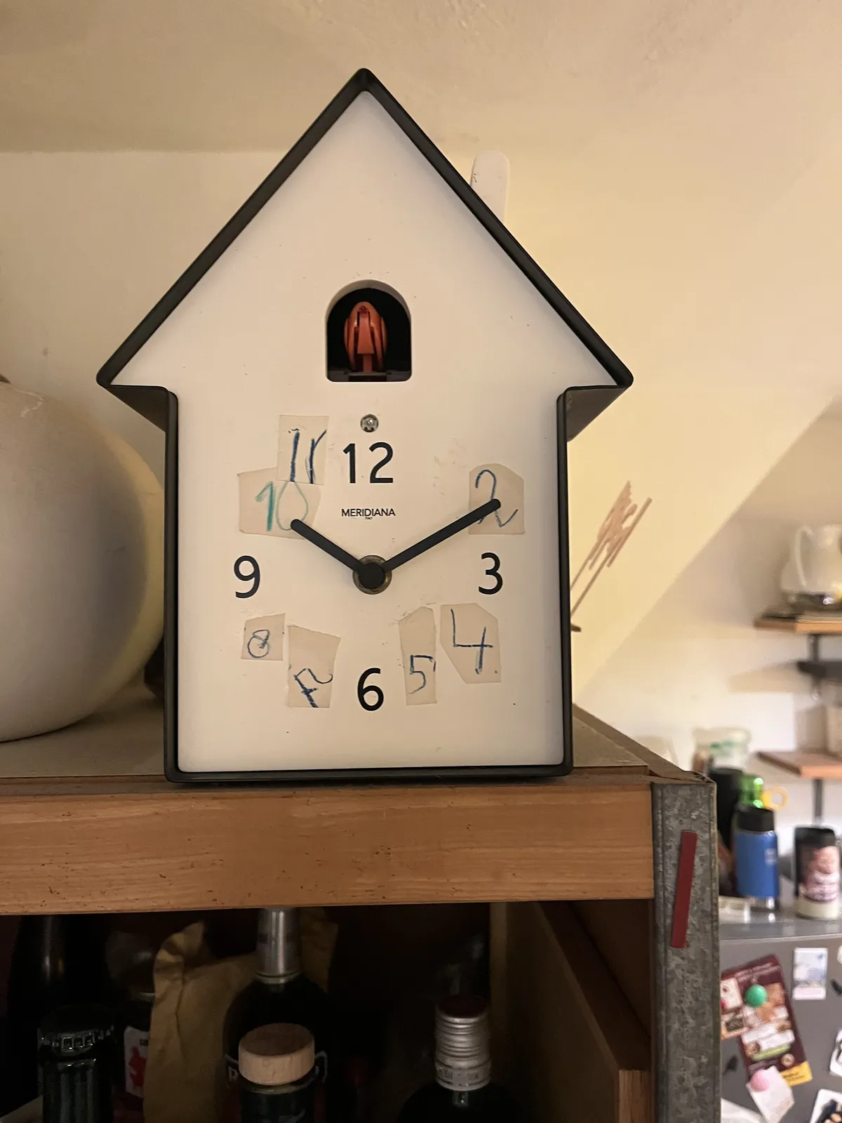 A modernist, minimalist cuckoo’s clock: geometrical, white with a black border. Numbers in children’s writing are pasted over the original numbers. The clock points at 10 past ten.