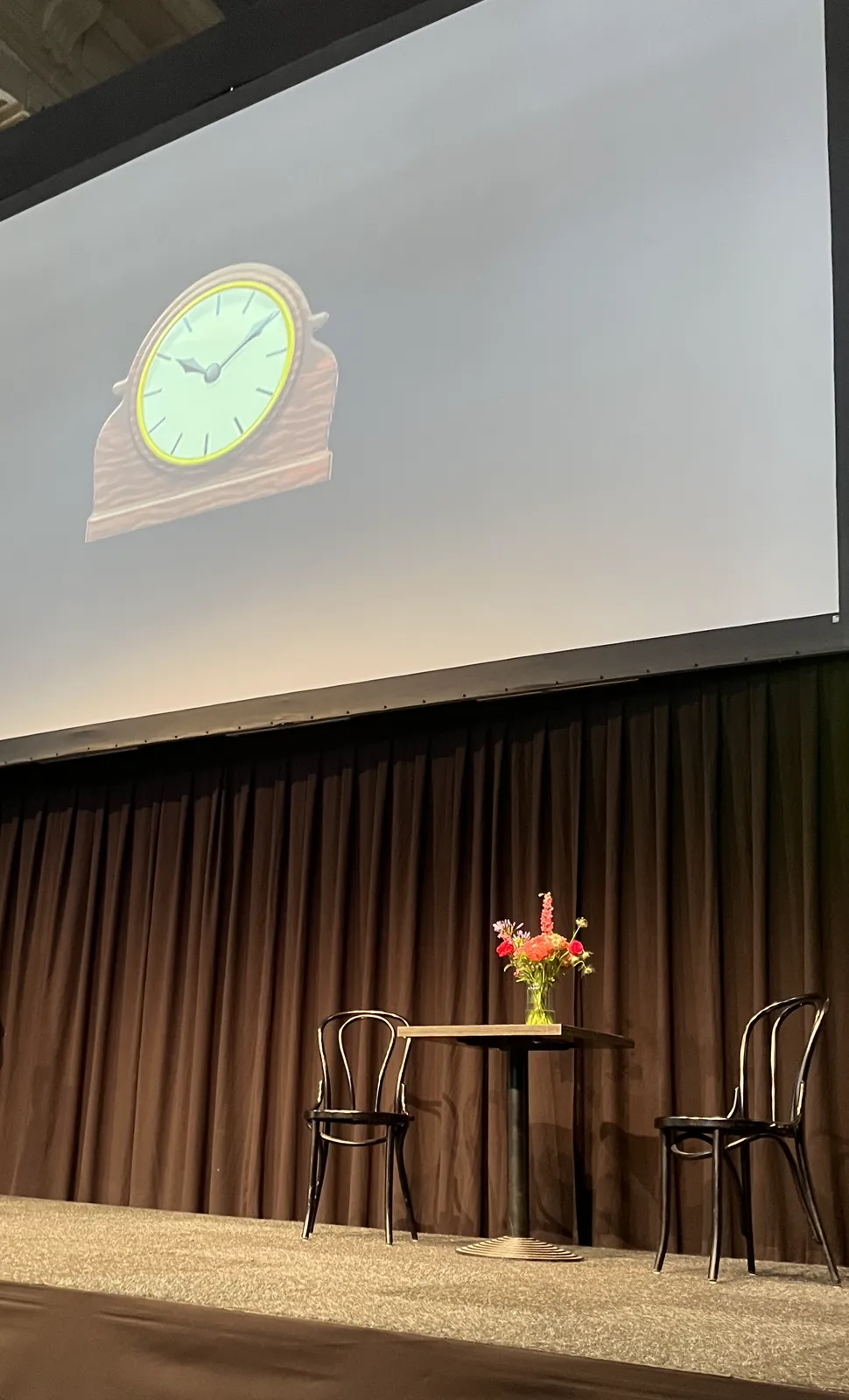 Two chairs on a stage, with a little table with flower on it between them. There’s a large screen behind the stage, with an emoji of a clock projected on it. This emoji points at ten past ten.