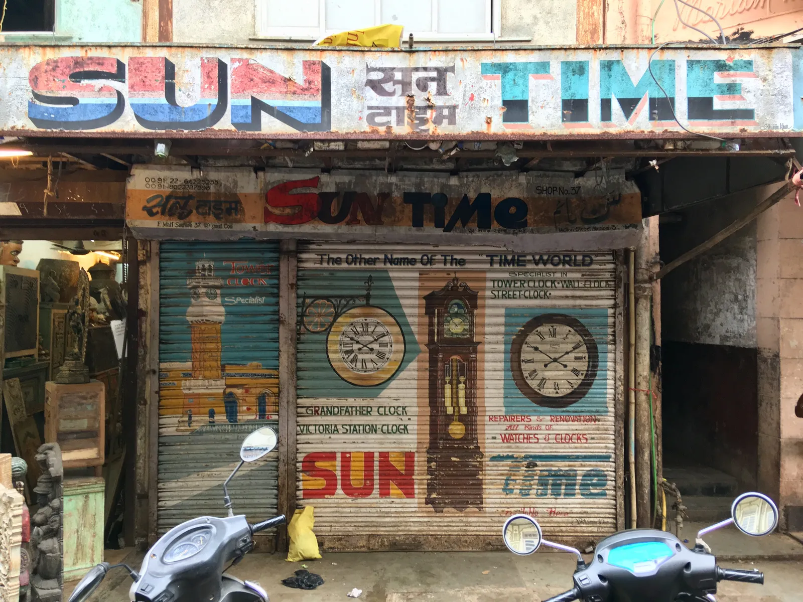 The shutter of Sun Time, a clock shop, is closed. Which is nice, because it has a few clocks painted on it. On two clocks the time can be clearly seen. They both point at 11 minutes past 10. There’s also the names of different types of clocks painted on the shutter. Grandfather Clock, Victoria Station Clock, Tower, Clock, Wall Clock, Street Clock. Sun Time, repairers and renovation, all kinds of watches and clocks.