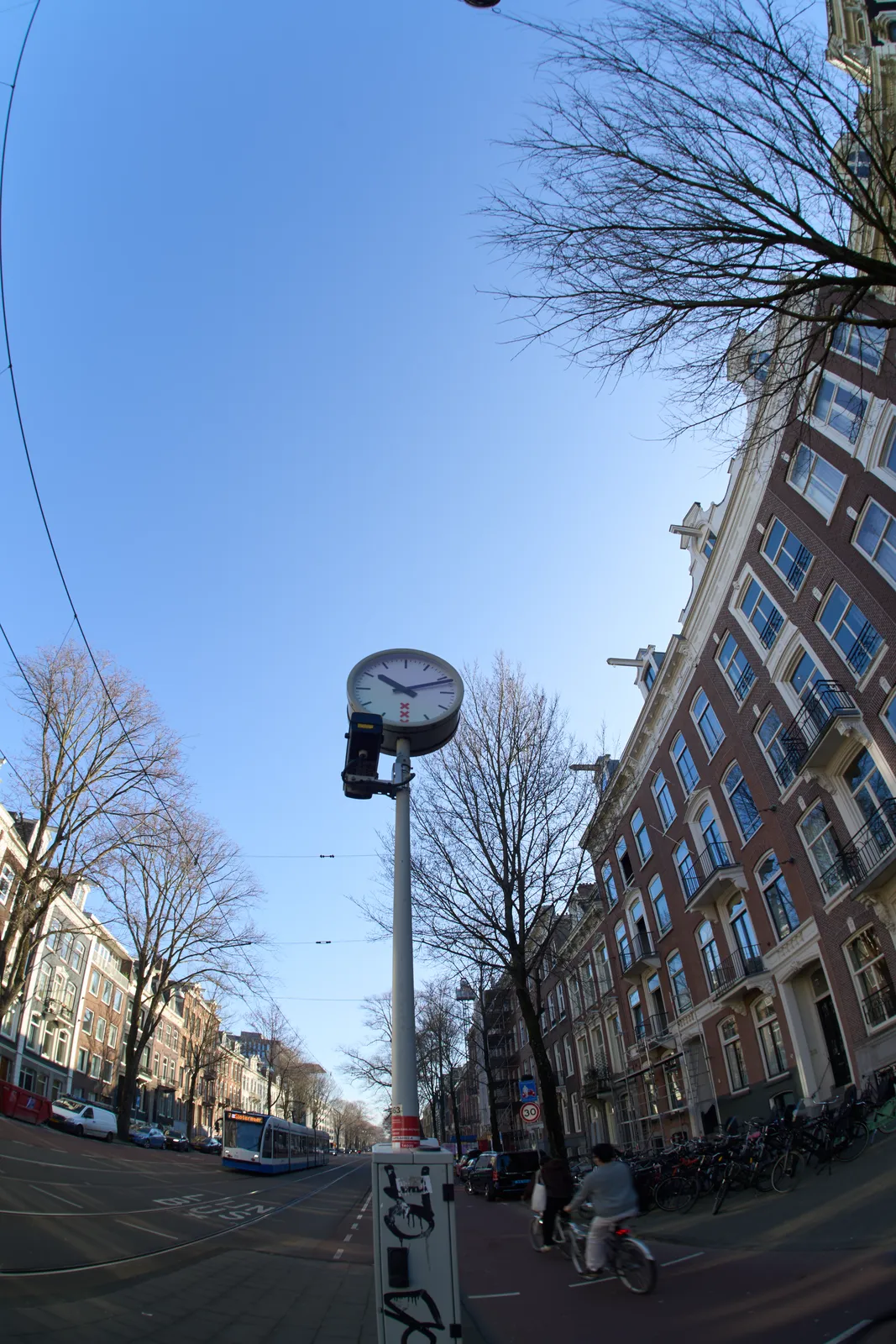 A very wide angle picture of a clock on a pole in an Amsterdam street. The clock points at 11 minutes past 10.