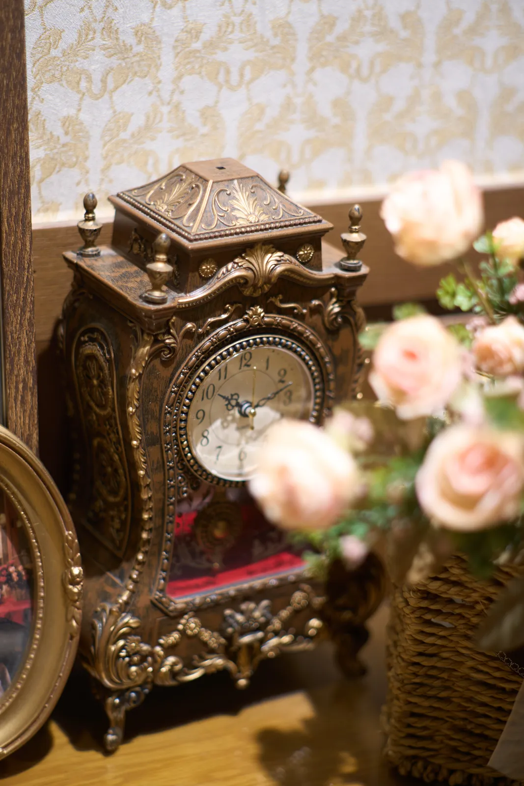A little, ornamented wooden desk clock, standing behind some flowers. It points at 12 minutes past 10.