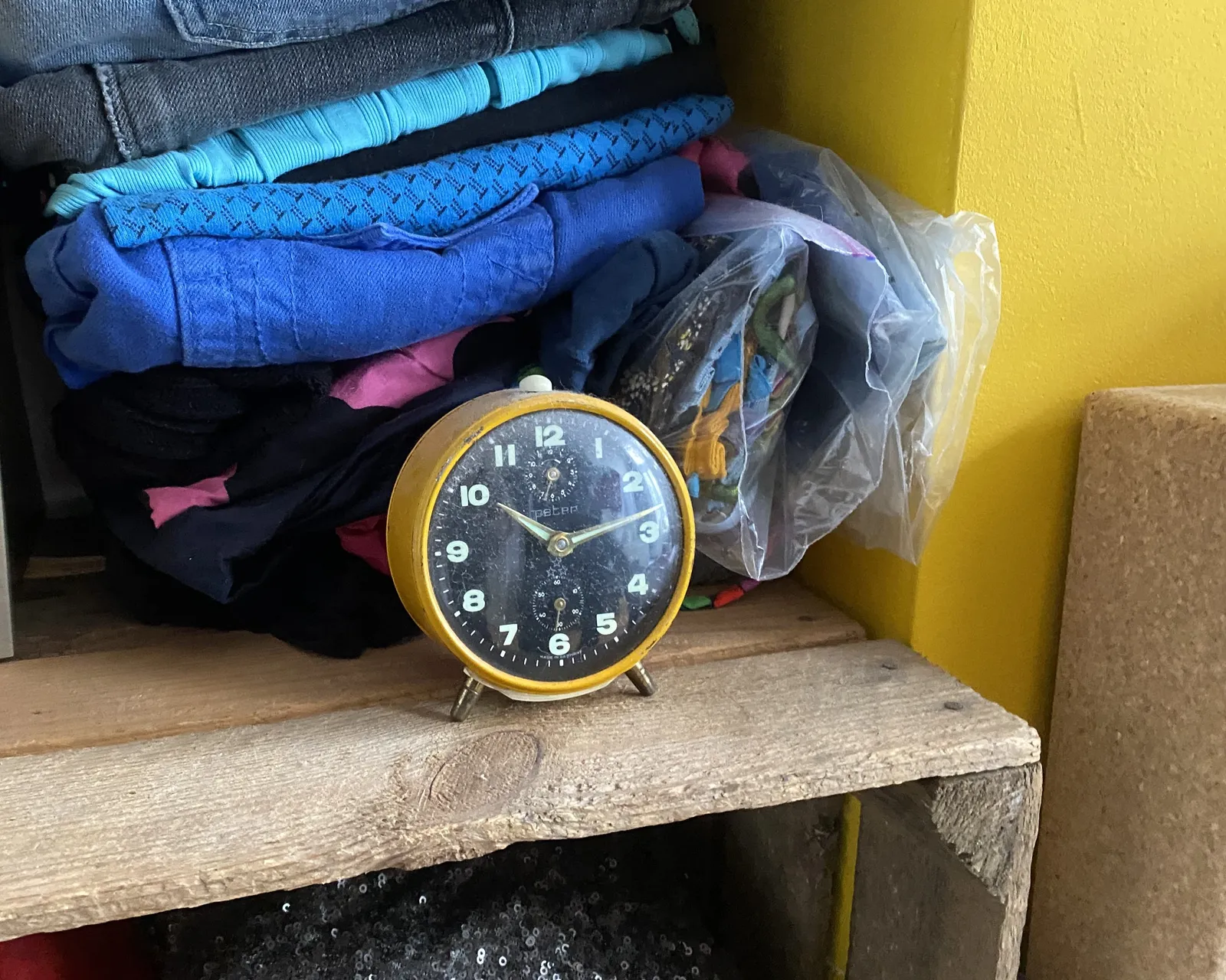 An alarm clock on a shelf in front of some clothes. It points at 13 past 10.