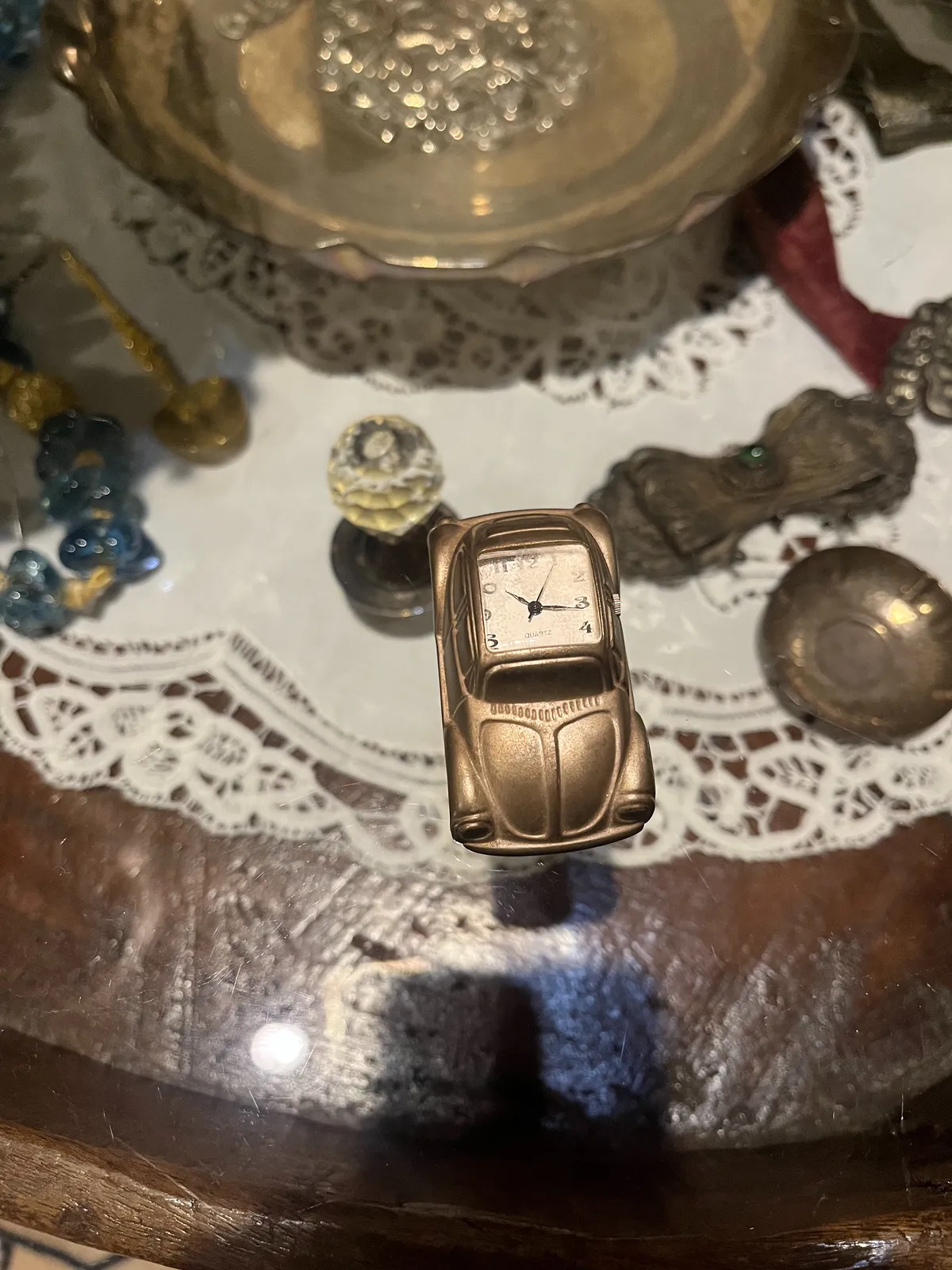 A little clock in the shape of an old Volkswagen Beetle standing on a glass table, with all kinds of silver items below it. The clock on the Beetle points at 16 minutes past 10.