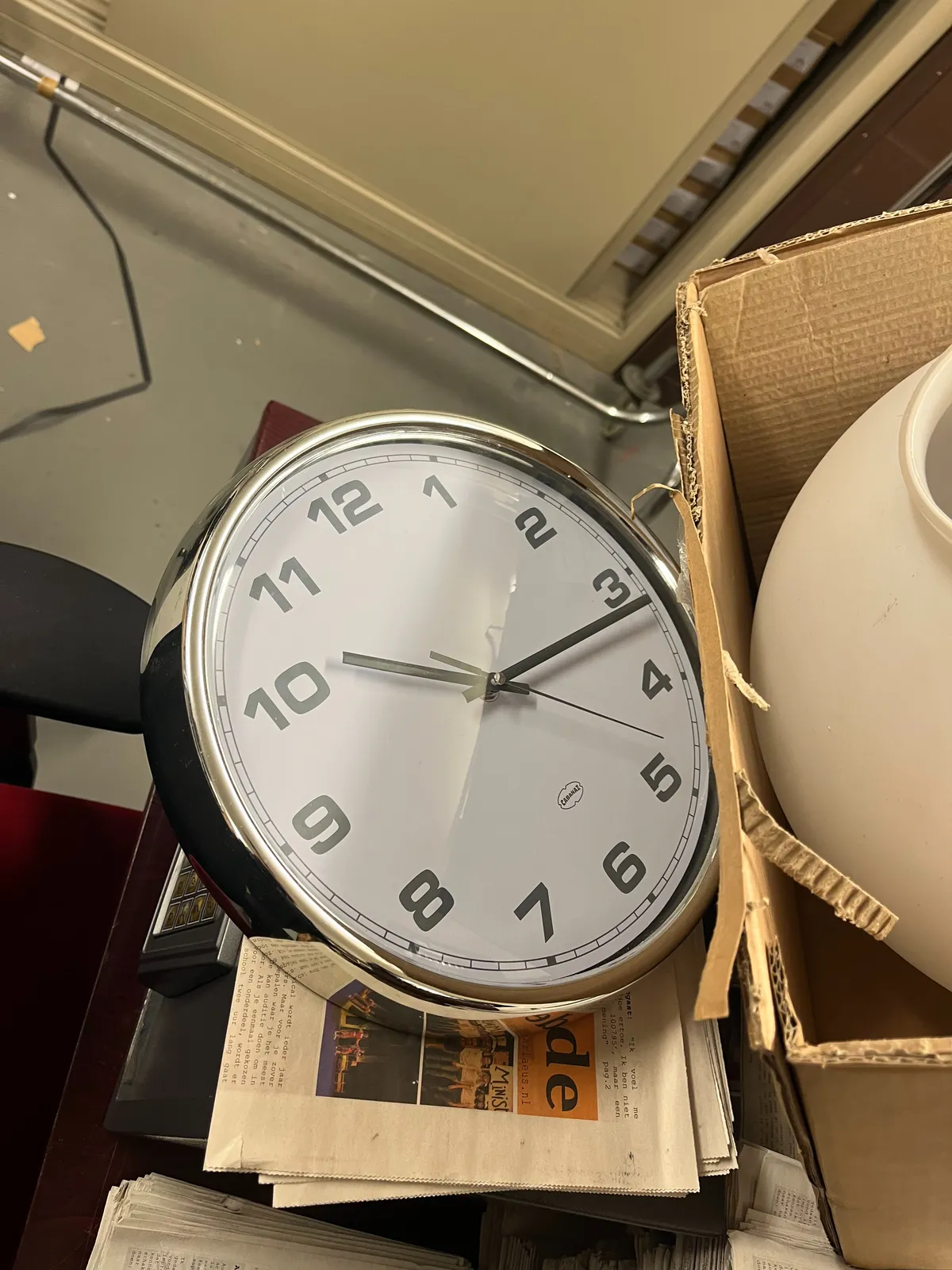 A clock laying on a pile of stuff. Its front glass is broken, yet the time can easily be read: It points at 16 minutes past 10.