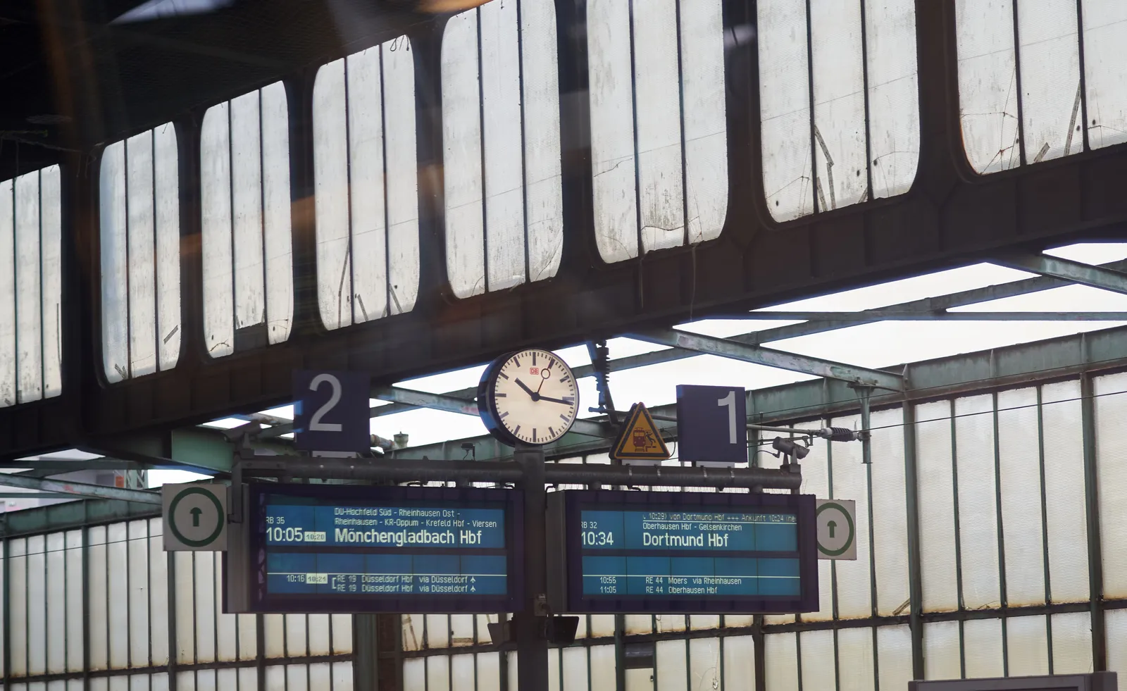 A clock on a train station, between two signs. The one sign gives information about the train to Mönchengladbach, the other about the train to Dortmund. On the clock it is 16 minutes past 10.