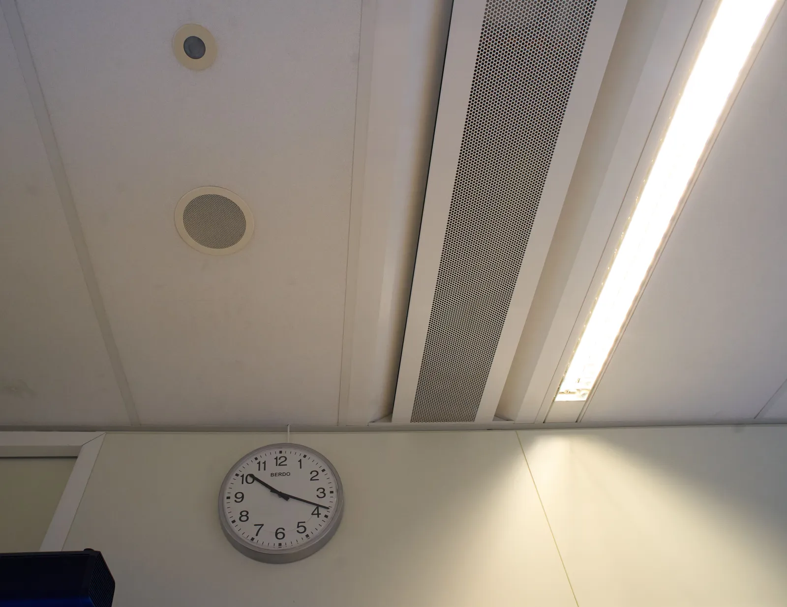 Looking a the upper part of a wall and a ceiling, inside a building that’s built from prefabricated system parts. There’s a clock on the wall that points at 18 minutes past 10.