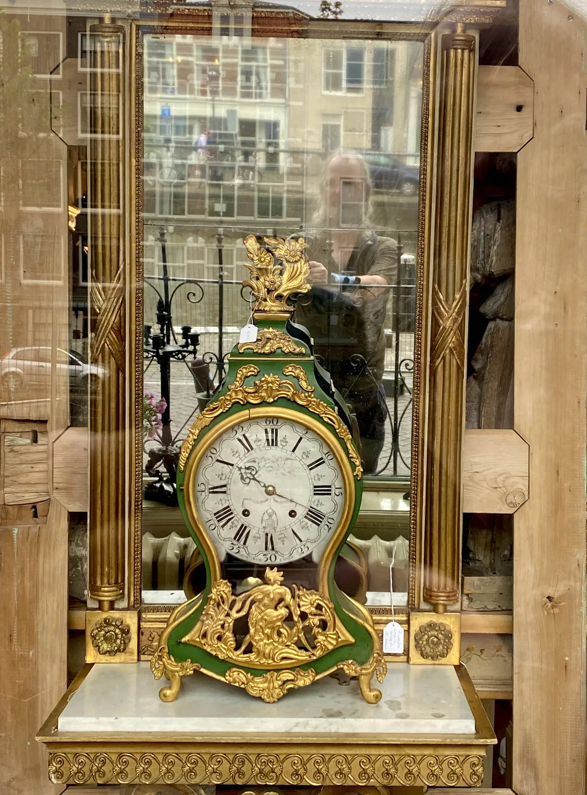 A richly ornamented, gold and green clock in front of an antique mirror. The photographer can be seen in this mirror. The clock points at 19 minutes past 10.