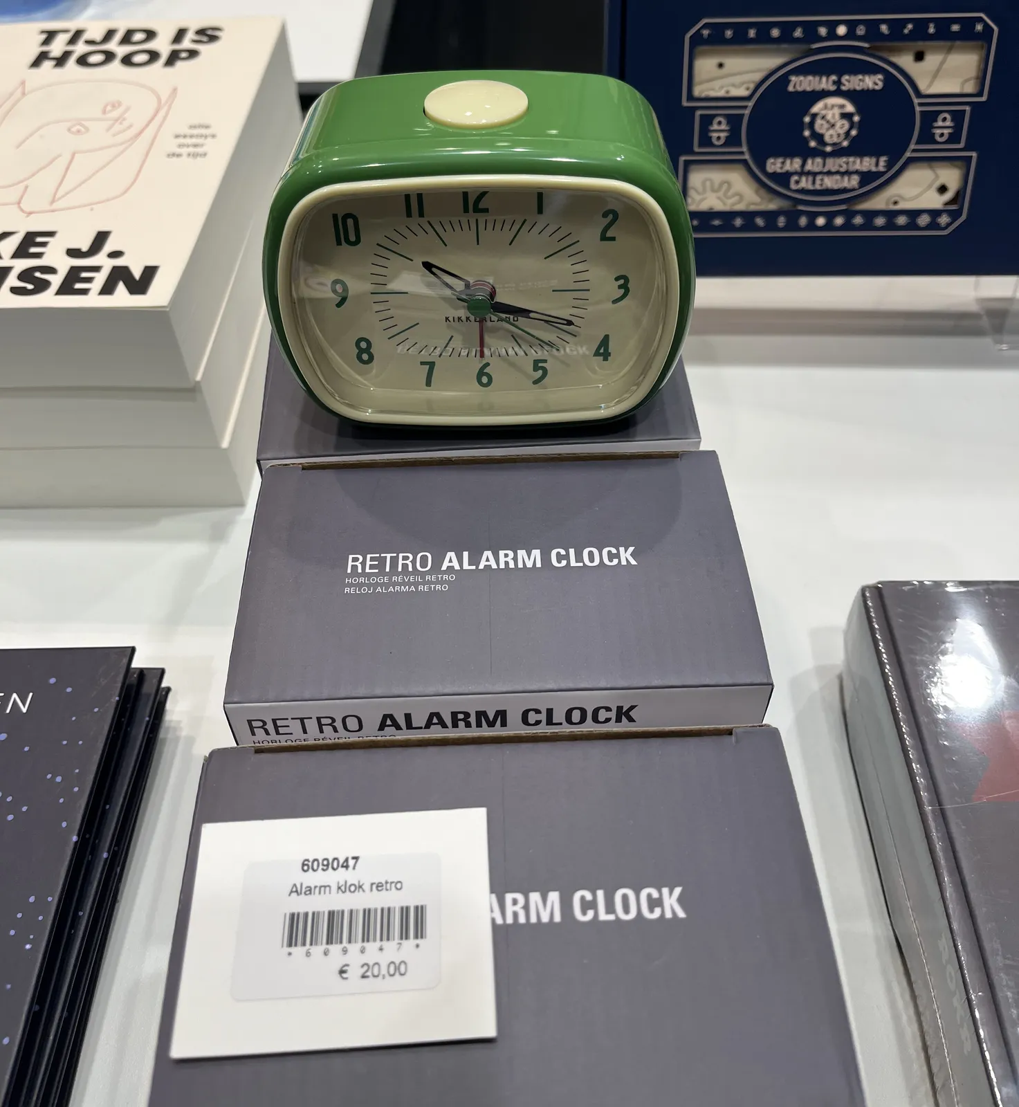 A green with pale white plastic alarm clock on top of a few boxes that say Retro Alarm Clock. The price is 20 euros. This retro alarm clock points at 19 minutes past 10.
