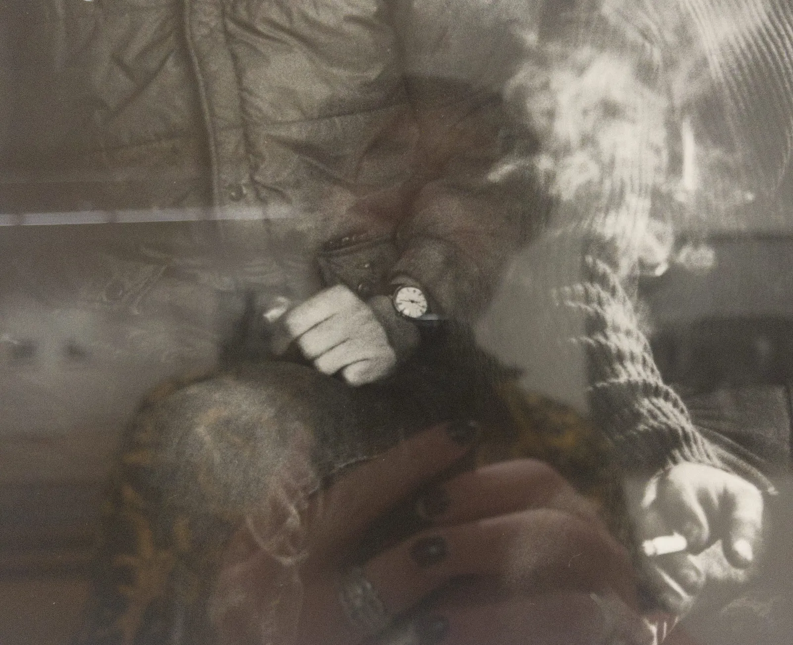 A picture of a part of a black and white picture of to people, and more specifically, the hands of two people. The hands of three people, if you count the hands of the person who takes the picture, who can be seen in the reflection. On of the people in the picture wears a watch over their sleeve. It poinst at 22 minutes past 10.