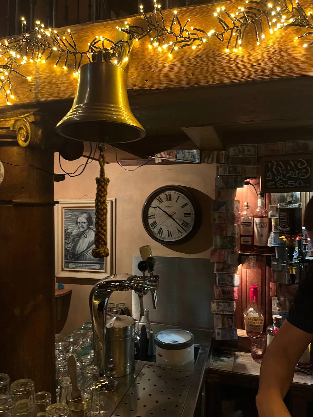 A big brass bell in the foreground, a tap, and a clock in the background in this pub. The clock points at 22 past 10.