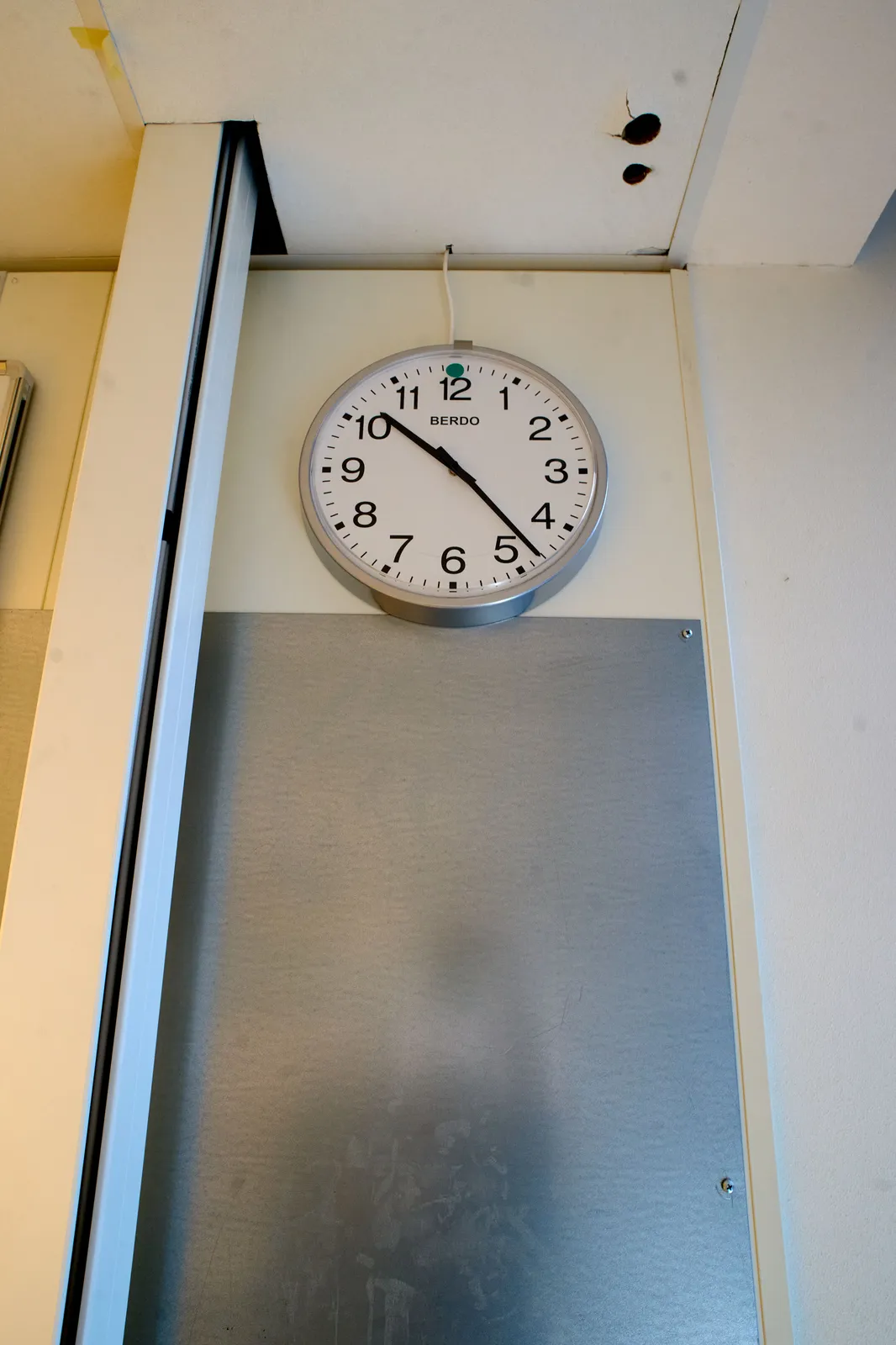 A metal sheet attached to a wall. A large cable duct runs down from the ceiling. There are two holes in the ceiling that might indicate that the duct was first placed a bit to the right. Not much love was given to placing this duct. There’s also a clock attached to the wall. It points at 23 minutes past 10.