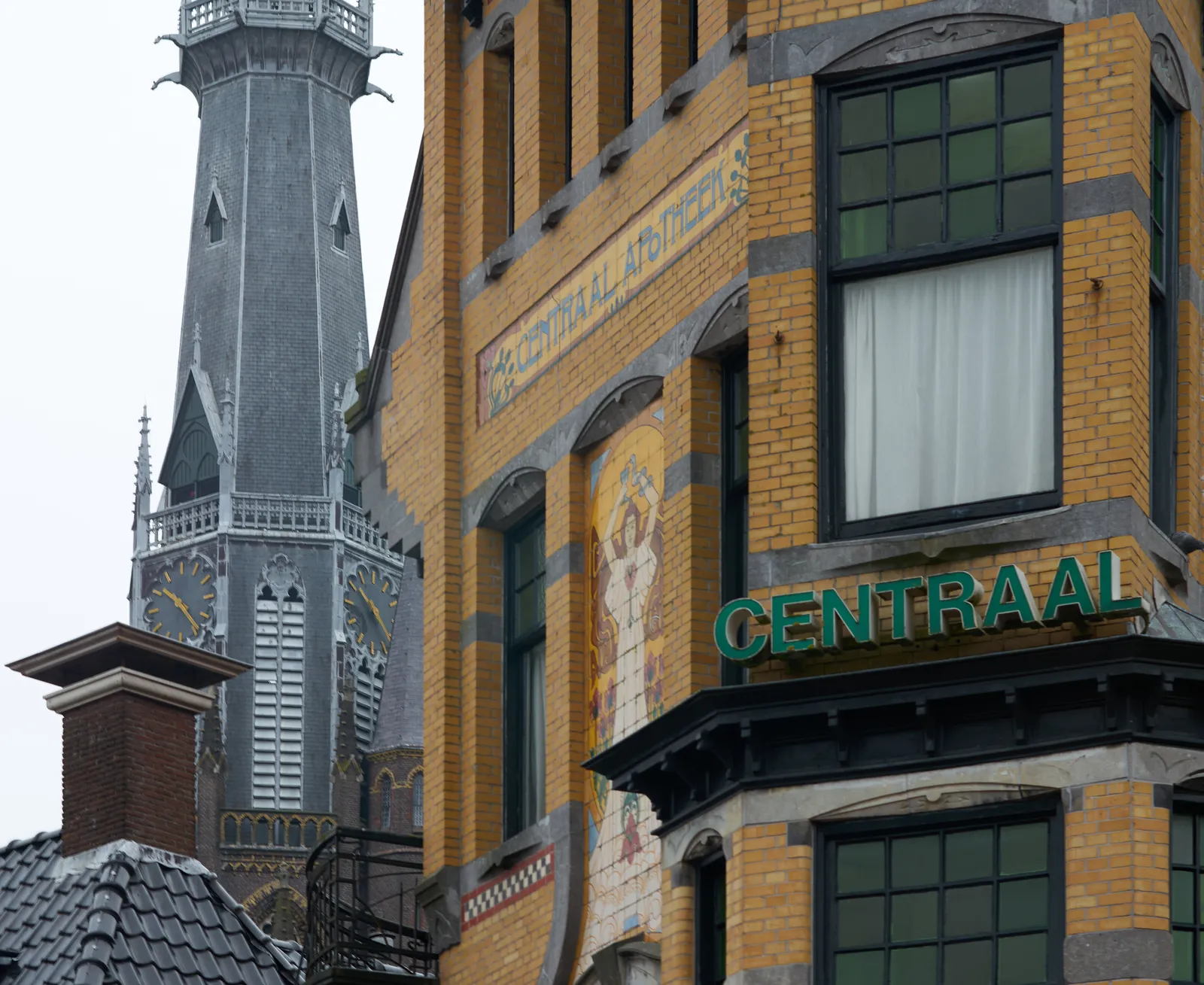 There’s a large church tower behind a yellow brick building. On the building are some art deco illustrations and the words Centraal Apotheek. On the church tower the clock points at 24 past ten.