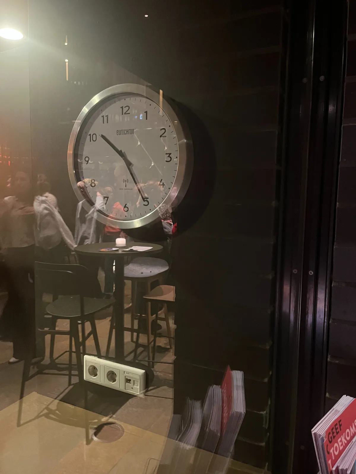 A clock behind glass. In the reflection you can see some people. The clock points at 25 minutes past 10.