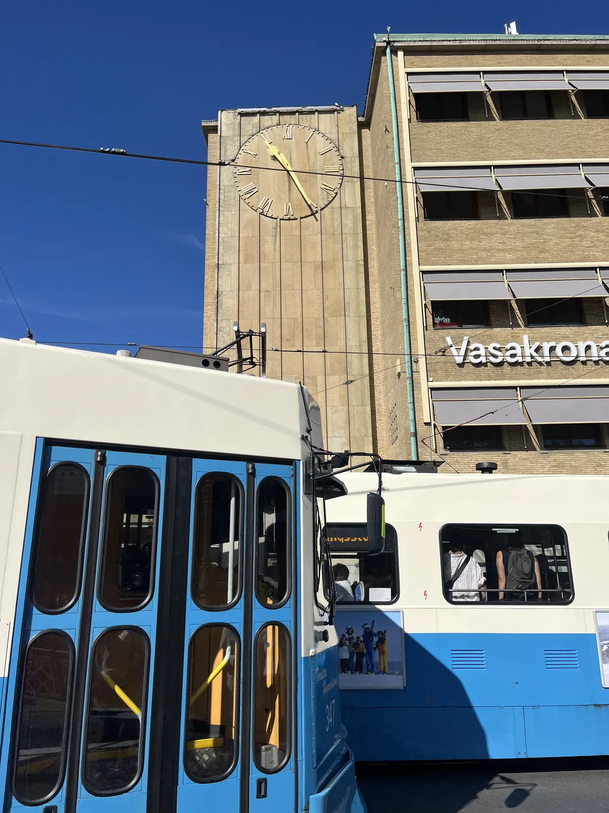Two blue trams pass each other in front of an almost brutalist building on a bright sunny day. On the building there’s a clock that points at 25 minutes past ten.