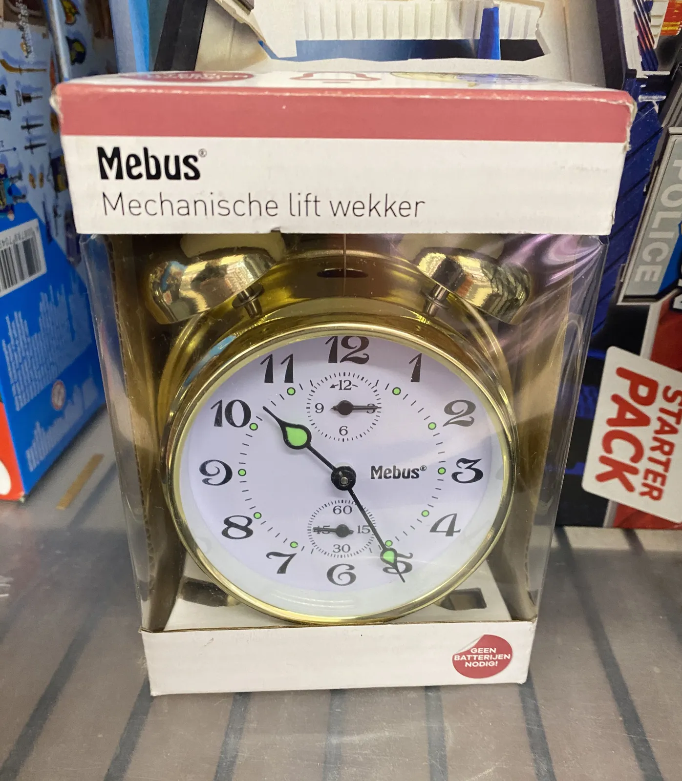 An old fashioned mechanical alarm clock in its original box. It says it is a mechanical lift clock. It points at 25 minutes past 10.