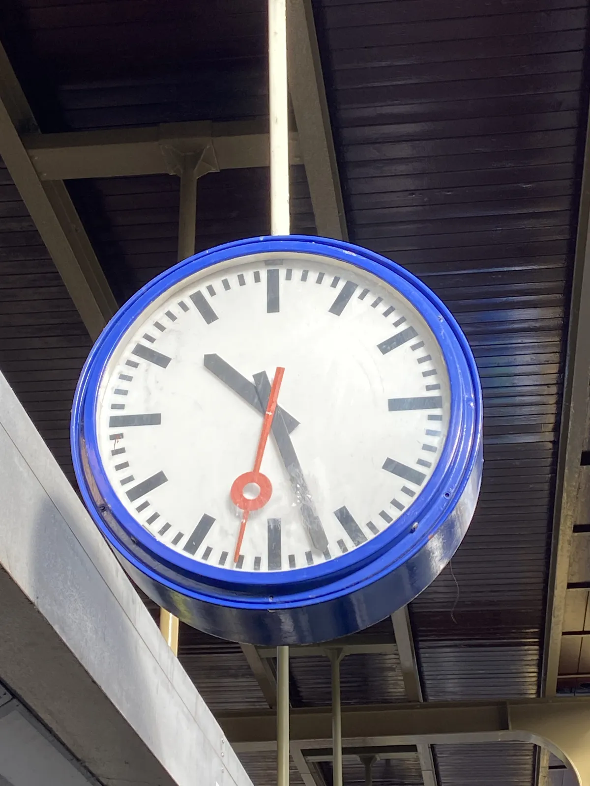 A clock attached to the ceiling of a train station. On it it is 27 past 10.