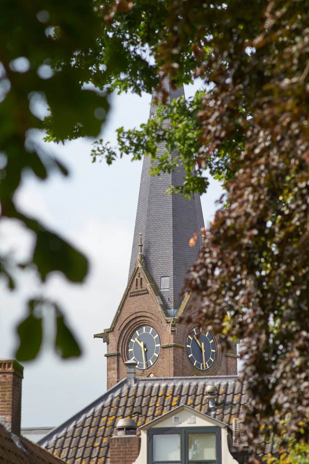 Behind some trees, behind a house, there’s a large old church tower with clocks on it. One of the clocks points at 28 past 10.