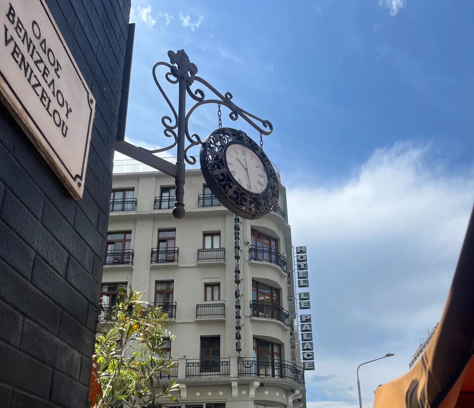 In the background the facade of Hotel Le Palace, in the foreground a road sign that says in Greek and latin letters that this is Venizelou road. Behind the sign there’s a clock attached to the corner of the building that points at 29 minutes past ten.
