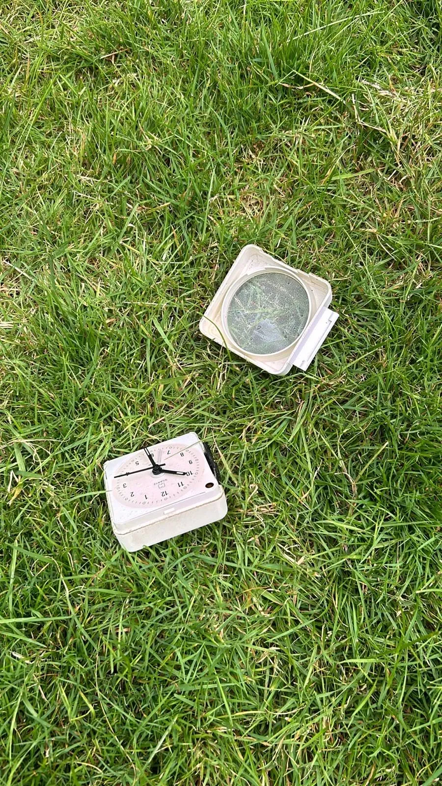 A broken, white alarm clock lies on the grass.  It seems like it was thrown out of the window at 29 minutes past 10.