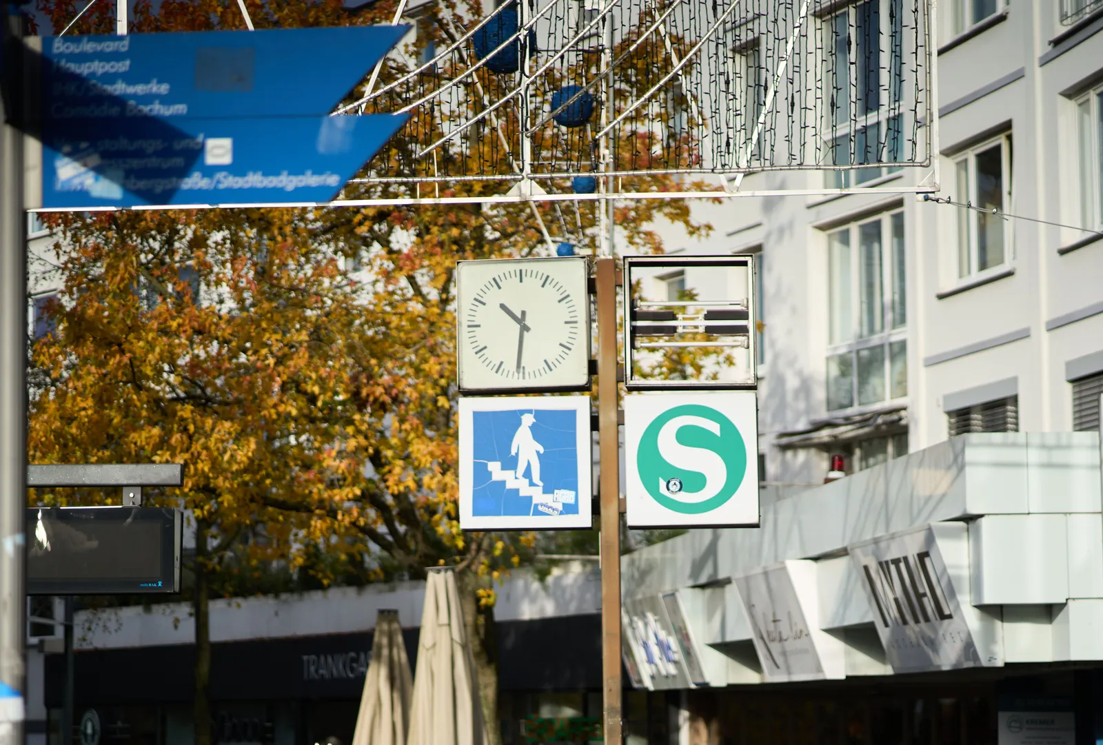 White appartment buildings in the background, and some signs, and a clock in the foreground. The clock points at 31 minutes past 10.