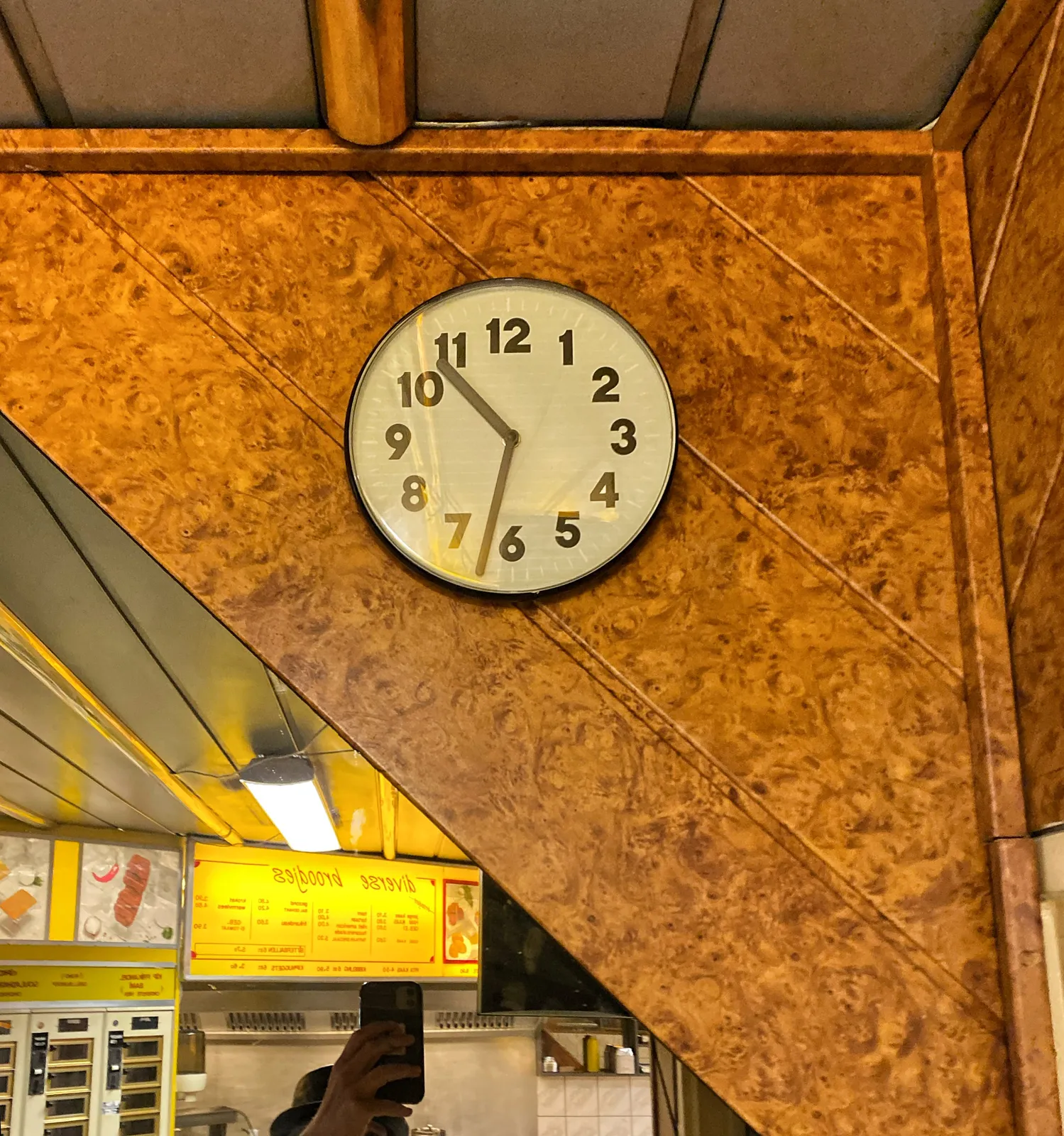 A clock in a snack bar, attached to the stairs. There’s also a mirror in which you can see the hand and the phone of the photographer. The clock points at 32 minutes past 10.