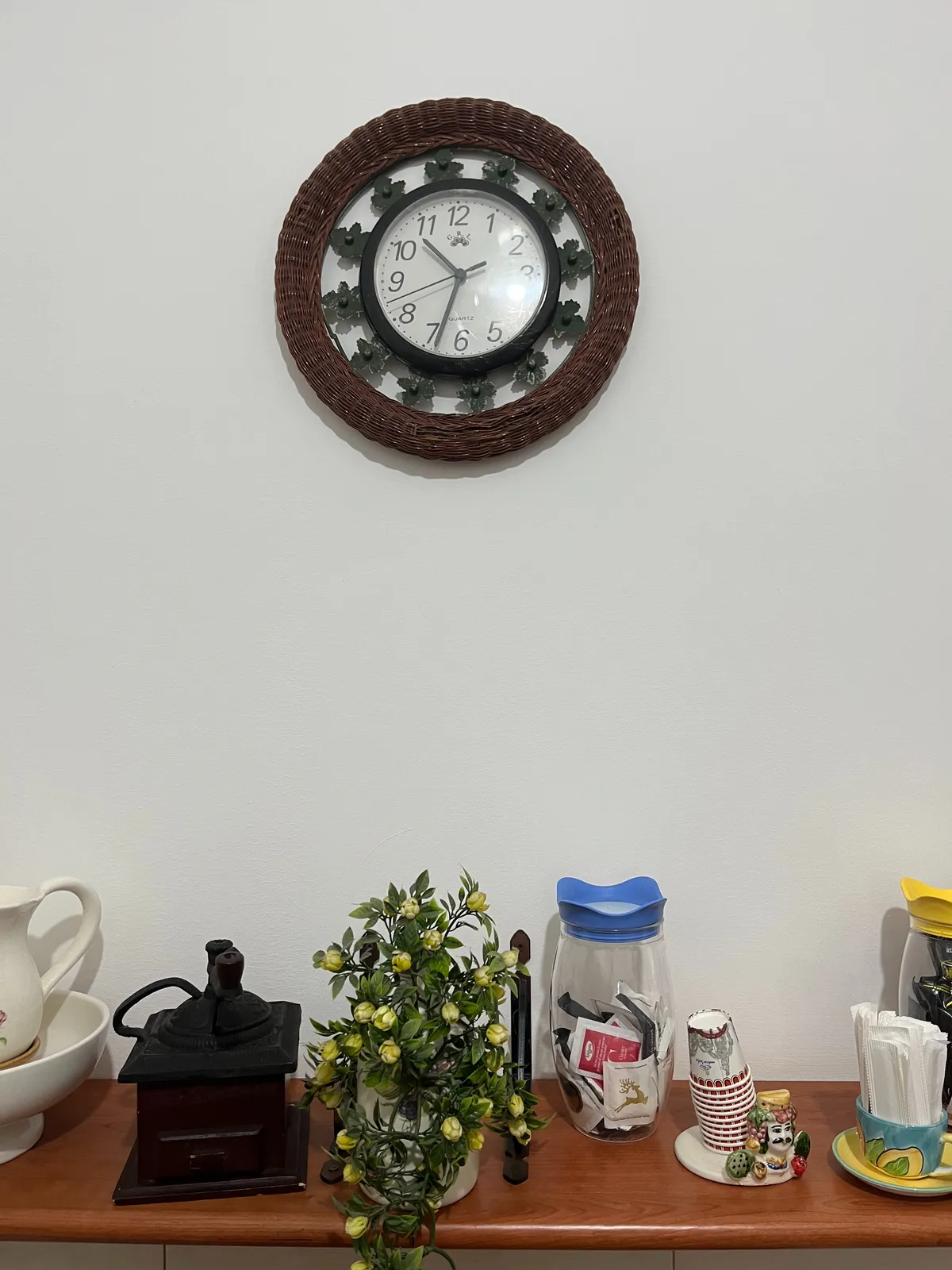 Some paper cups, some sugar, a plant and some other stuff on a shelf. Above this shelf there’s a clock on the wall which points at 33 minutes past 10.