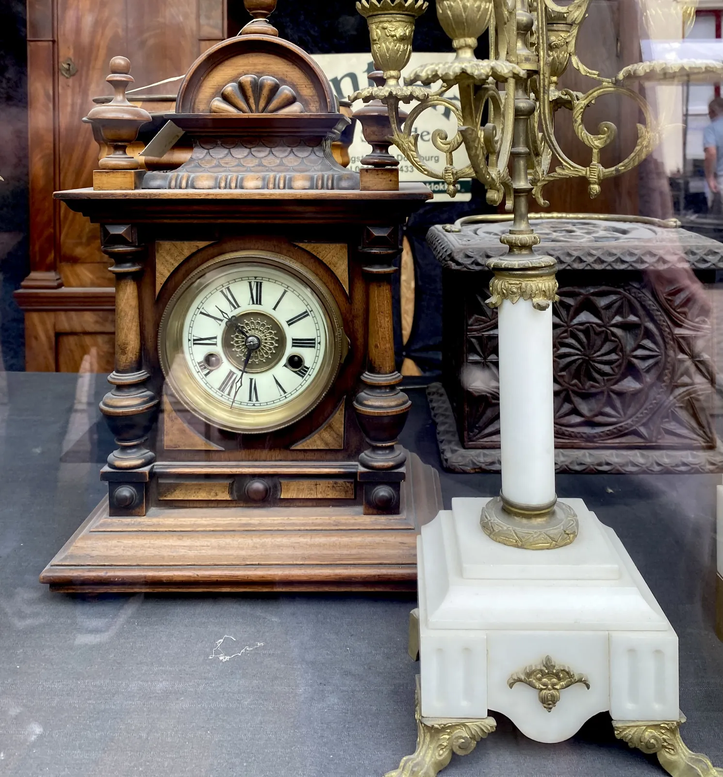 An antique wooden clock in a shopping window, among other antiques. It points at 33 minutes past 10.
