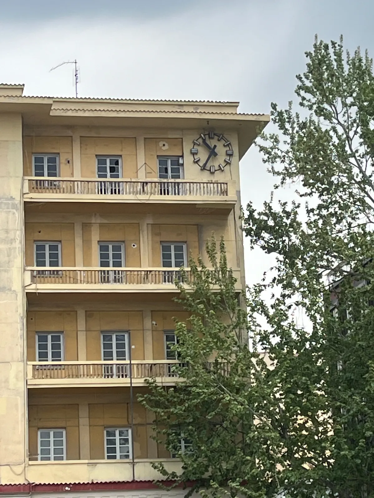 A clock on the wall of a balcony on the top floor of a yellow appartment building. It points at 34 minutes past ten.