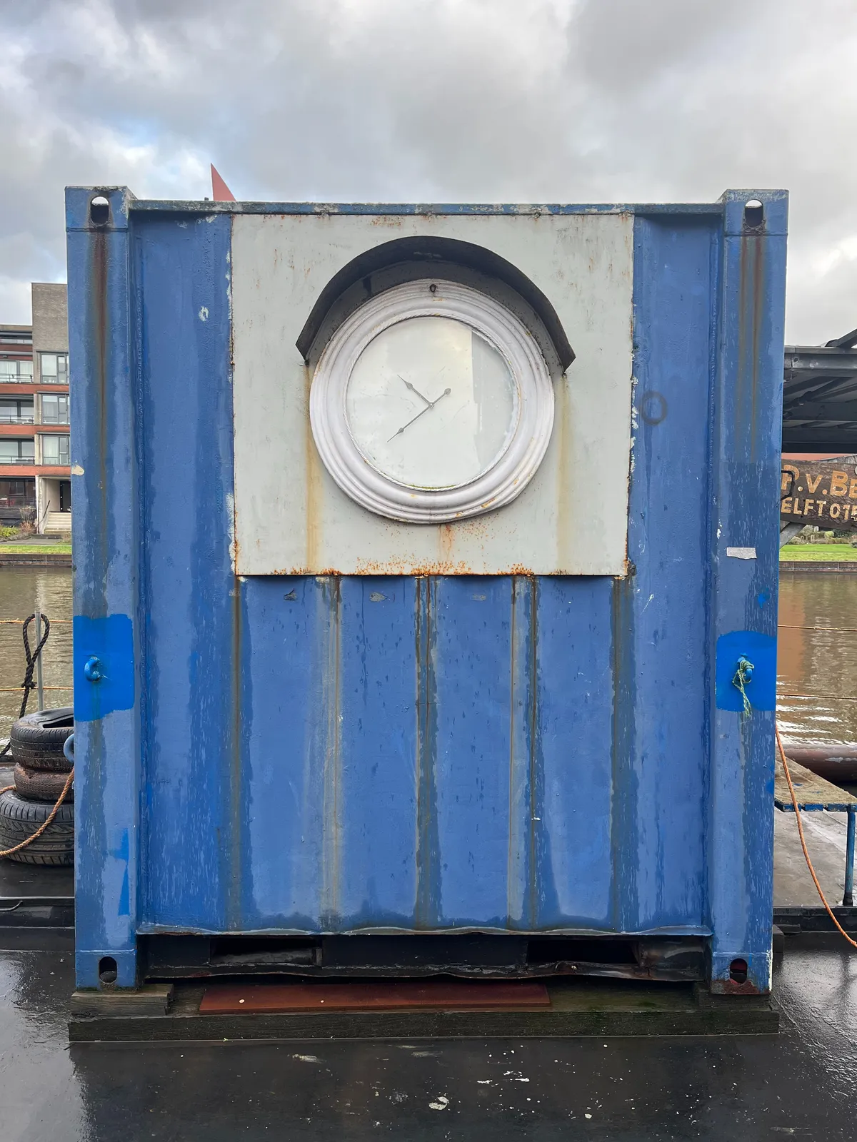 On a small blue container which seems to be standing on a boat there’s a clock that points at approximately 21 minutes to 11. 
