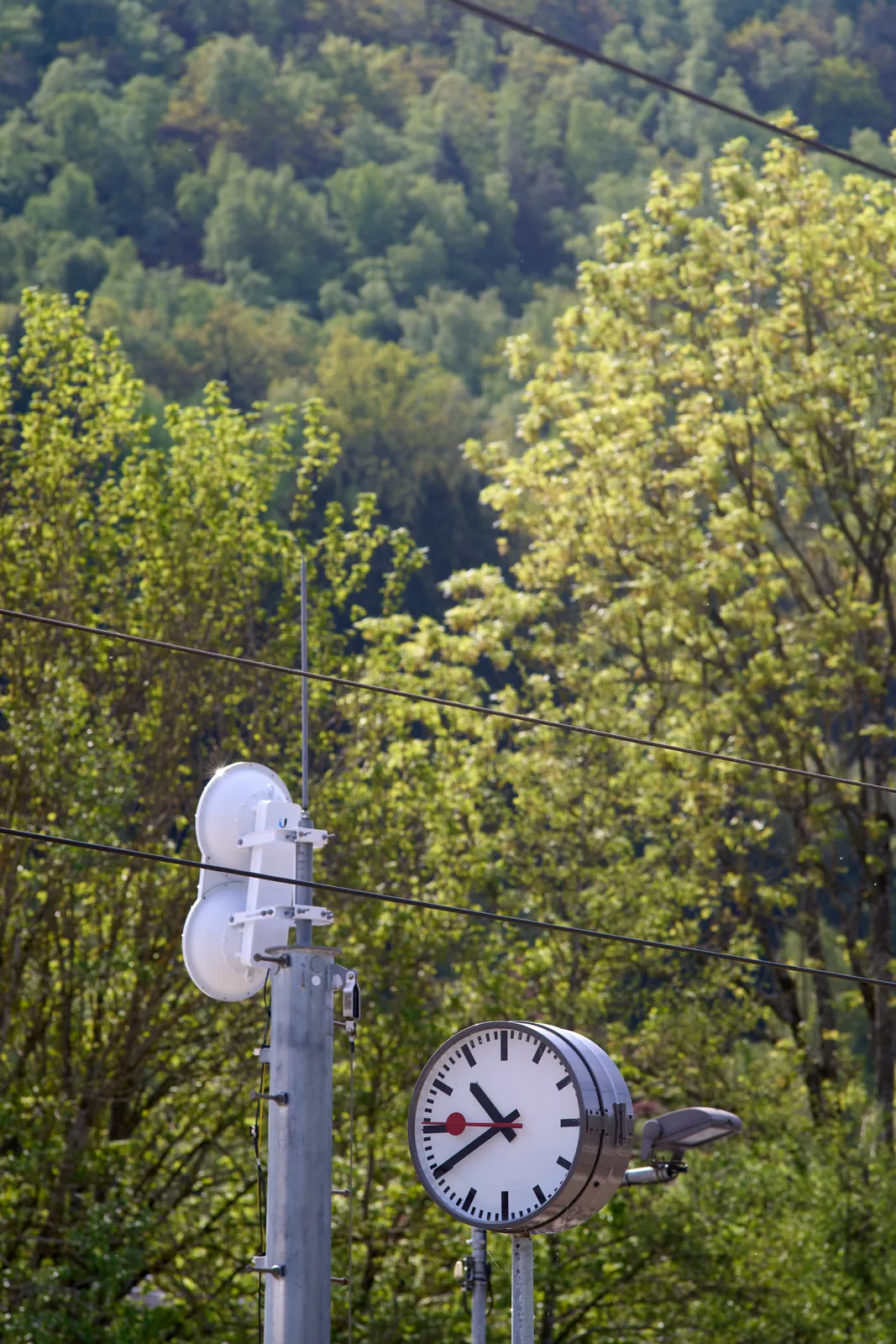 In the background there’s a mountain with a forest. In the foreground is a station clock which points at 20 minutes to eleven.