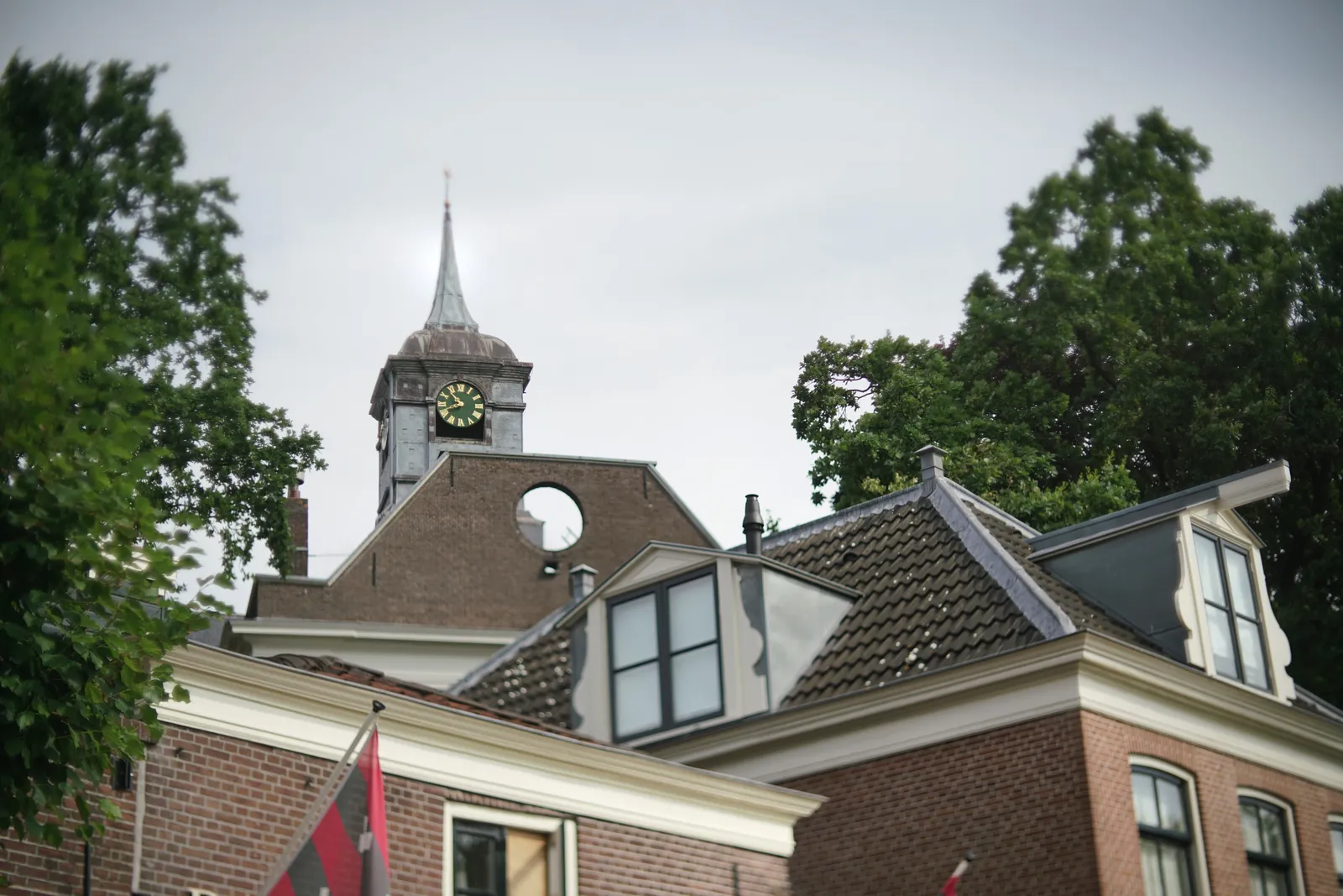 A composition of old buildings and trees. In the back there’s a clock, and in front of it there’s a wall with a circular hole in it. It looks like you should be able to see the clock through the hole, but unfortunately there’s a house on that exact viewpoint. The clock points at 18 minutes to 11.
