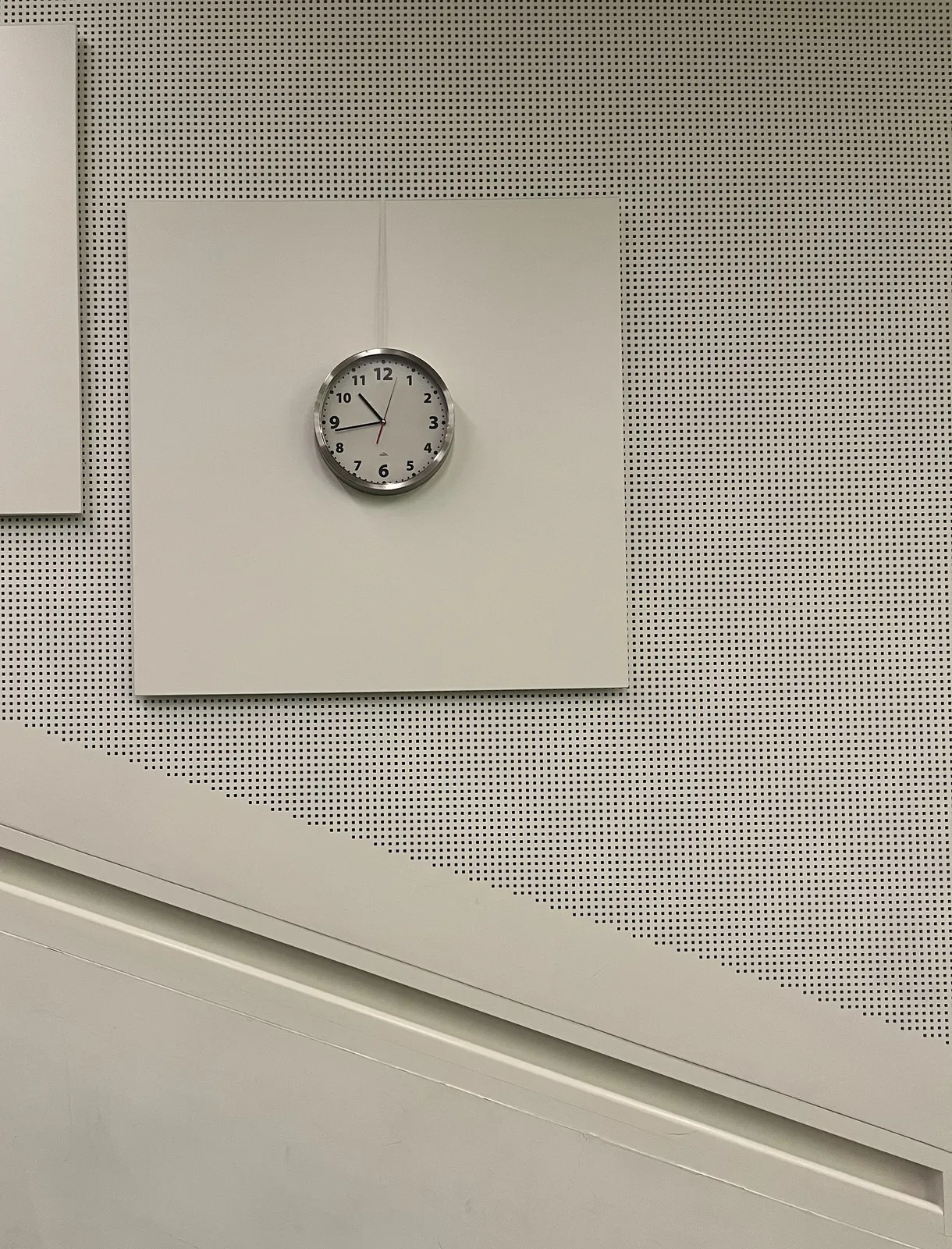 A white wall. There’s a diagonal in this wall. The upper part of the wall has a grid of small dots. There’s a white square on this grid with a clock on it. This clock points at 17 minutes to 11.