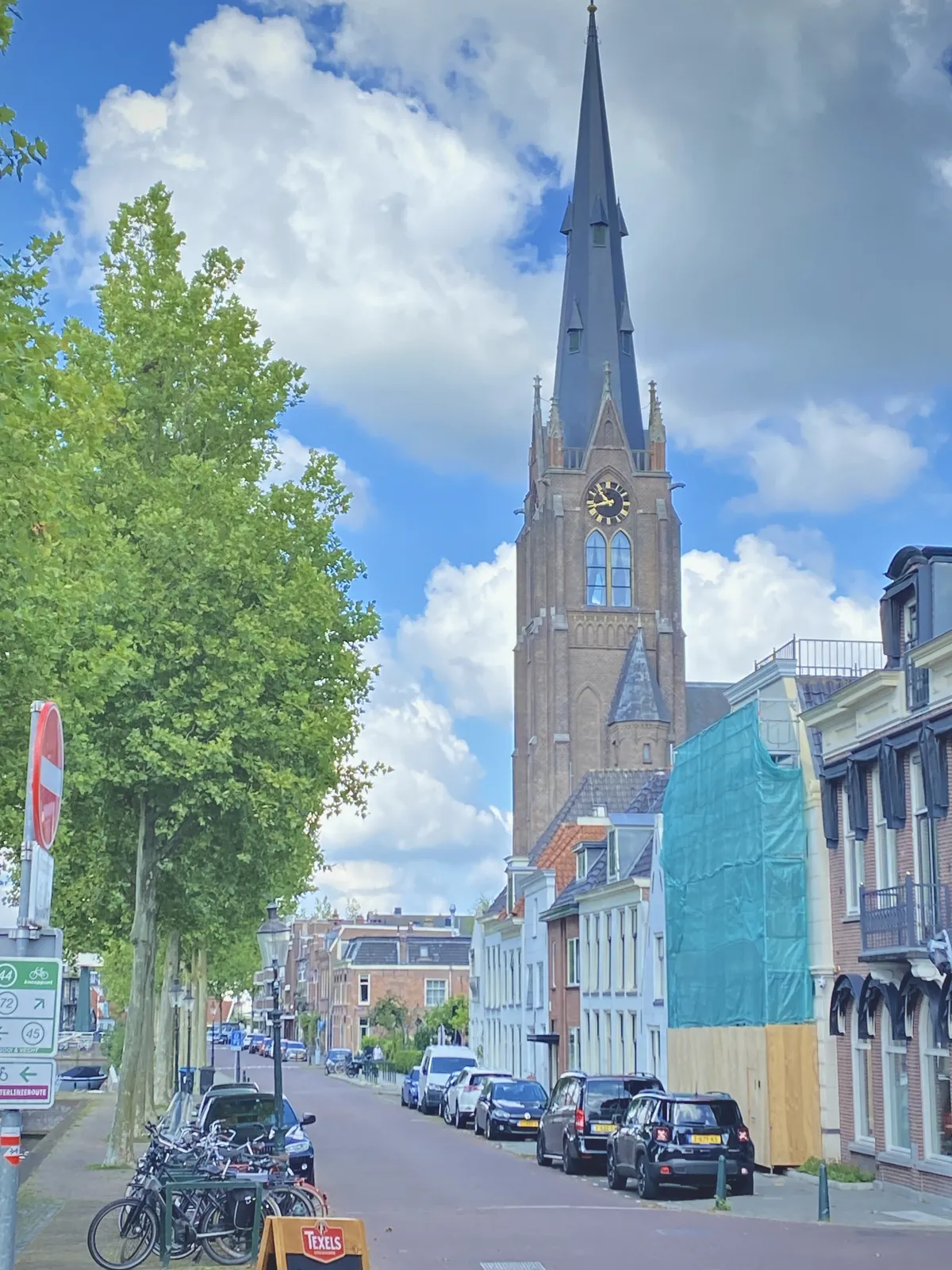 A Dutch street. Trees on the left, houses with parked cars in front of it, on the right. In the background there’s a huge church tower with a clock. It points at 17 minutes to 11.