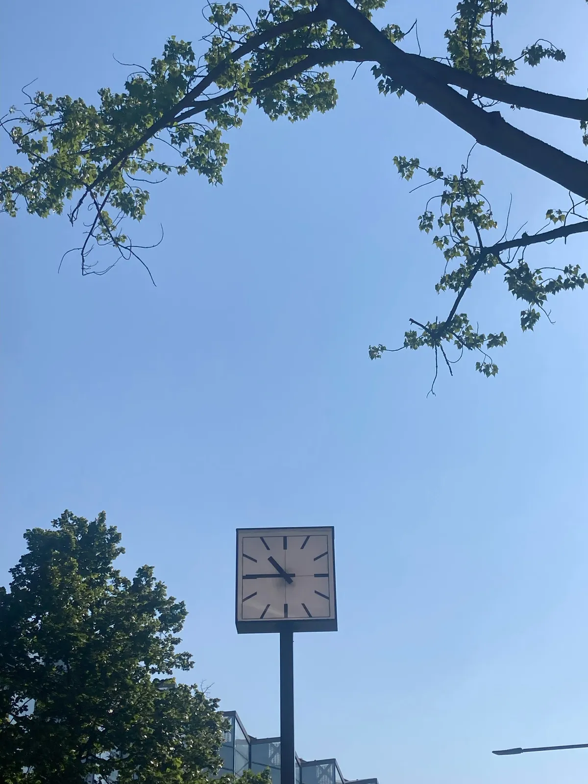 A square clock on a pole on a sunny day. It points at a quarter to eleven.