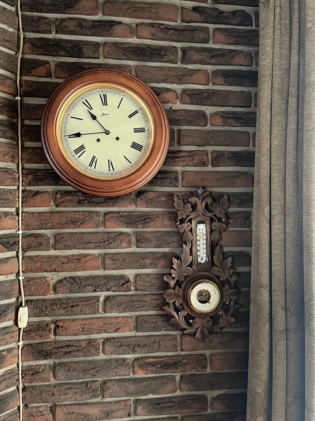 A wall clock on a brick wall, next to a richly ornamented, dark, wooden barometer. There’s a gray curtain on the right, and a white cord with a switch on the left. The clock points at a quarter to 11