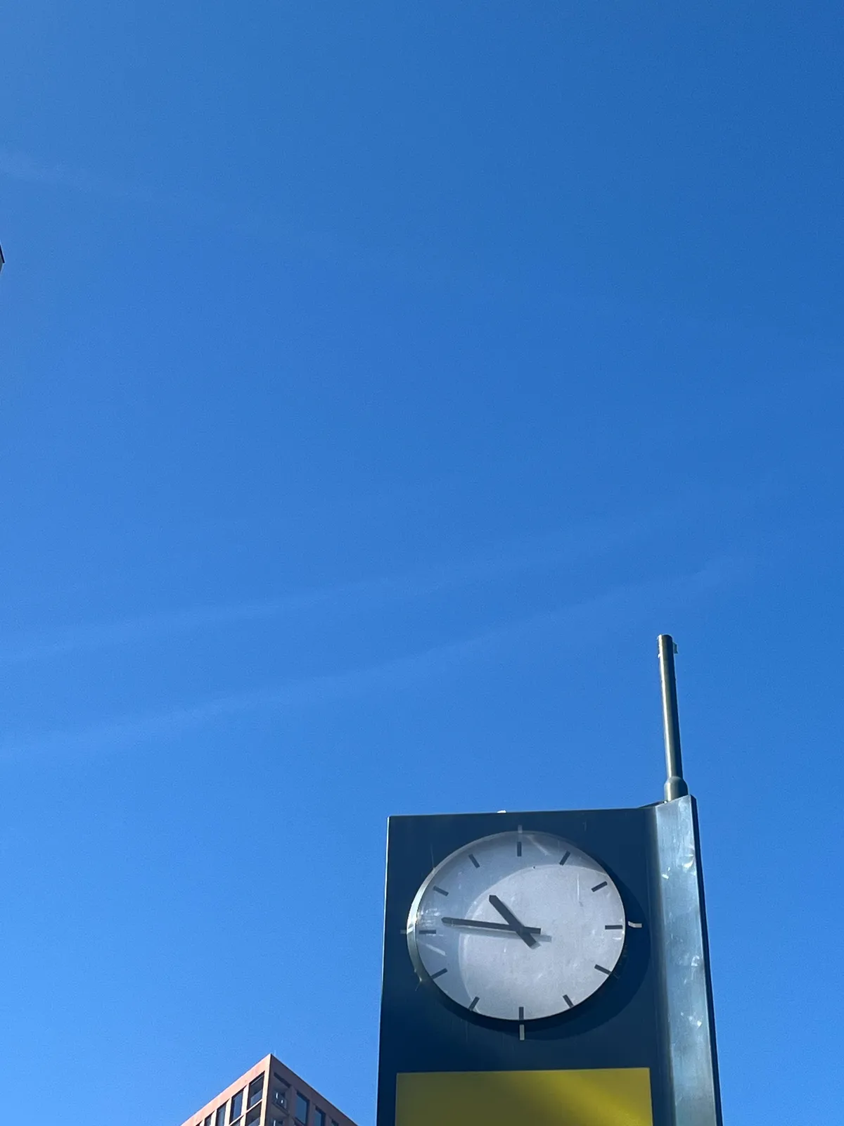 A bright blue sky. In the bottom left corner the top of a highrise can be seen. In the bottom right corner there’s a clock that points at 14 minutes to 11.