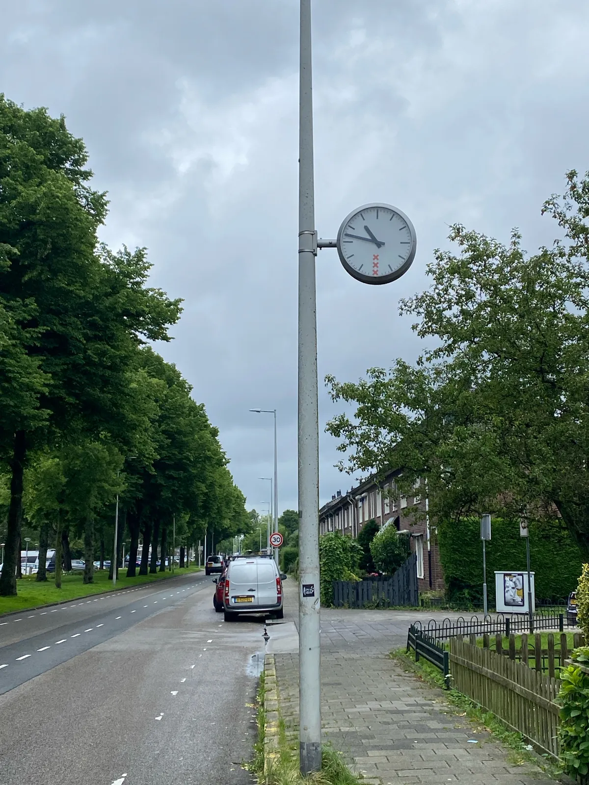 A clock with the Amsterdam Municipality logo on its face, attached to a pole in a street with trees on the left, and houses on the right. The clock points at 13 minutes to 11.